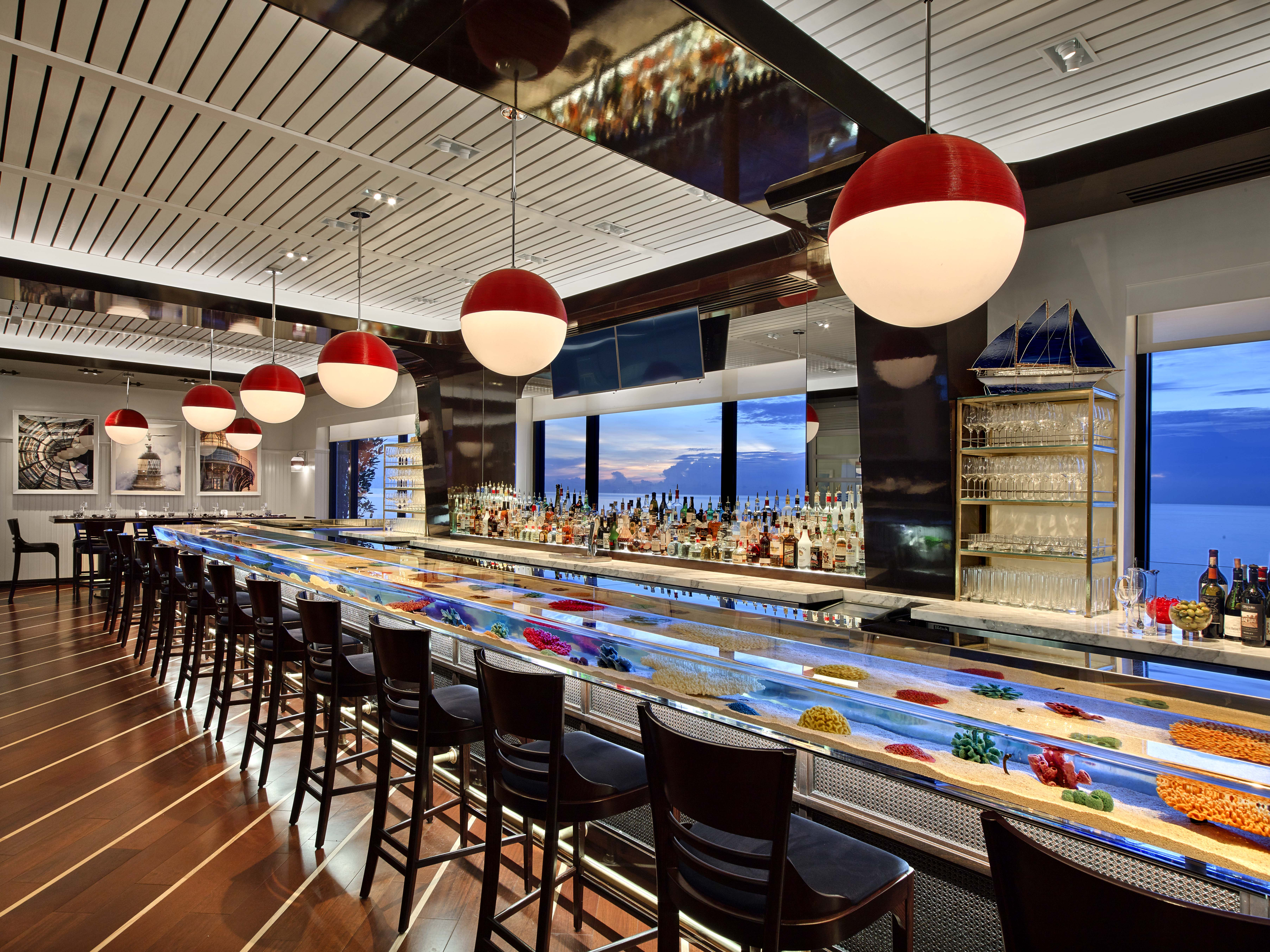 Modern bar with red pendant lights, illuminated counter with colorful display, and ocean view windows.