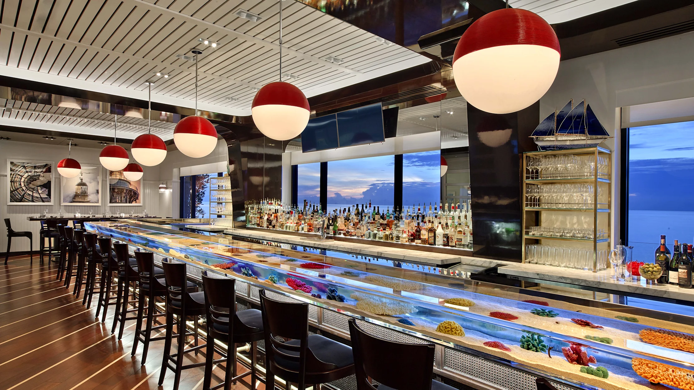 Modern bar with red pendant lights, illuminated counter with colorful display, and ocean view windows.