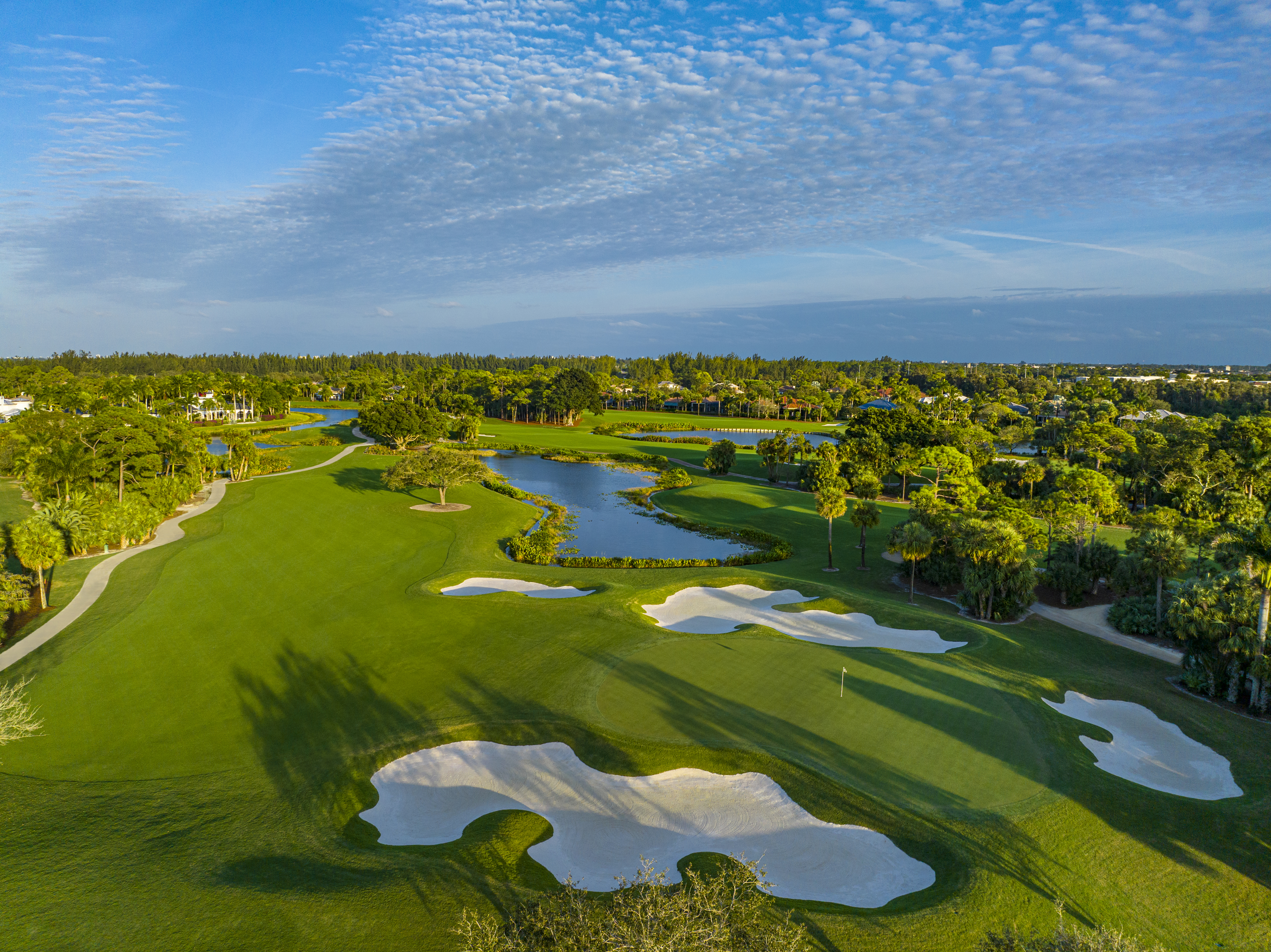 Aerial view of lush green golf course with white sand bunkers, water hazards, and blue sky with clouds.