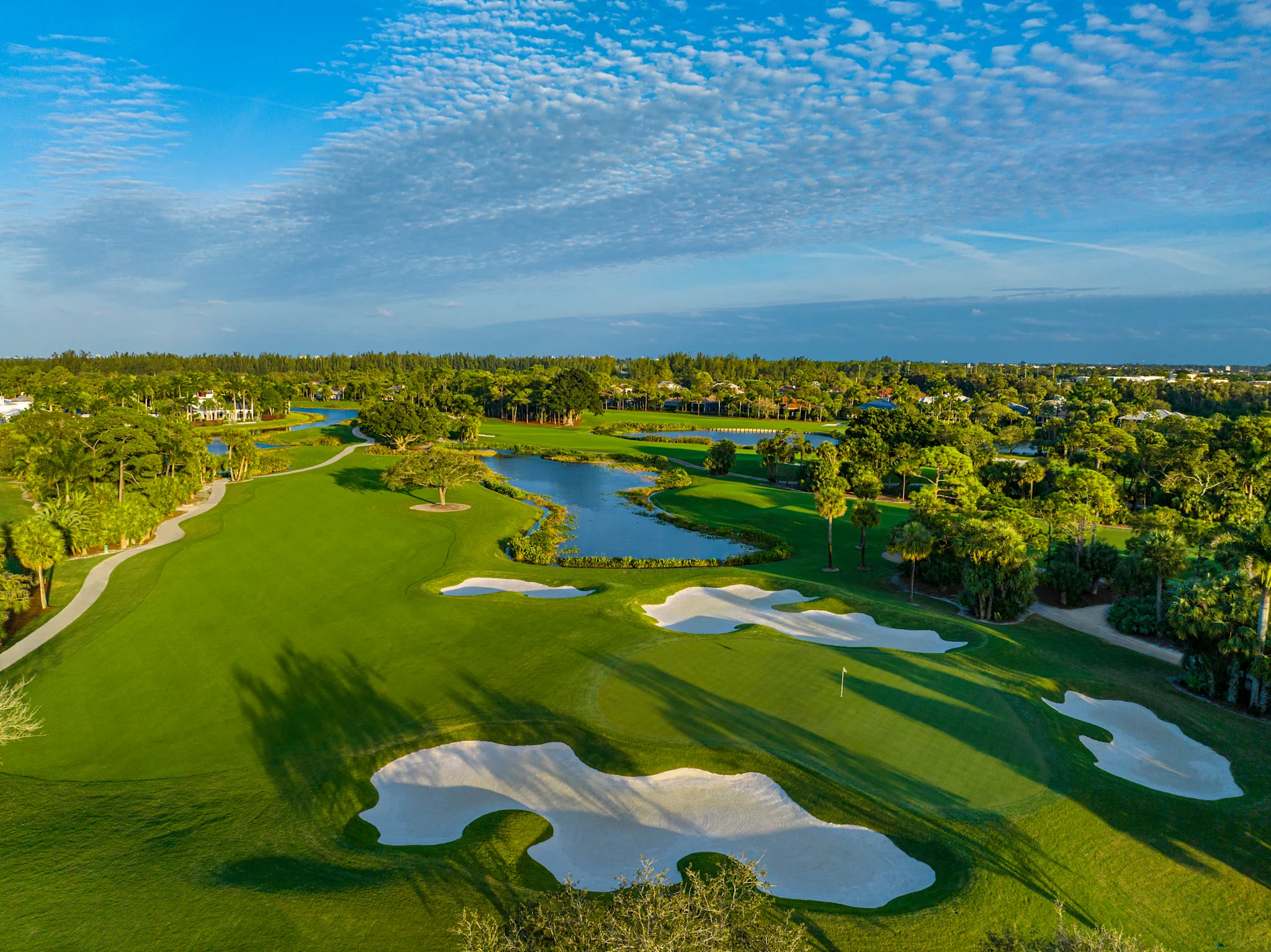 Aerial view of lush green golf course with white sand bunkers, water hazards, and blue sky with clouds.