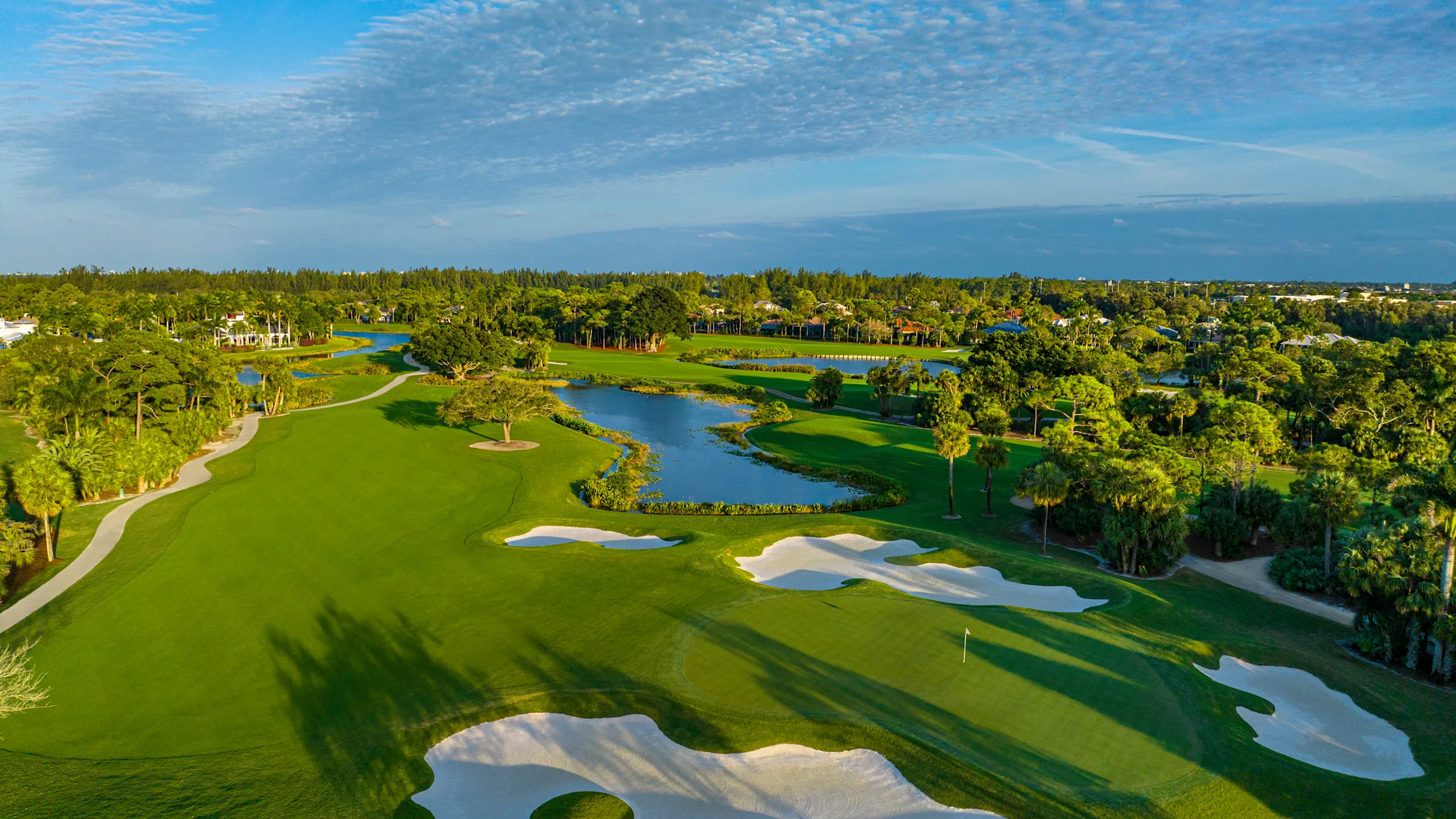 Aerial view of lush green golf course with white sand bunkers, water hazards, and blue sky with clouds.