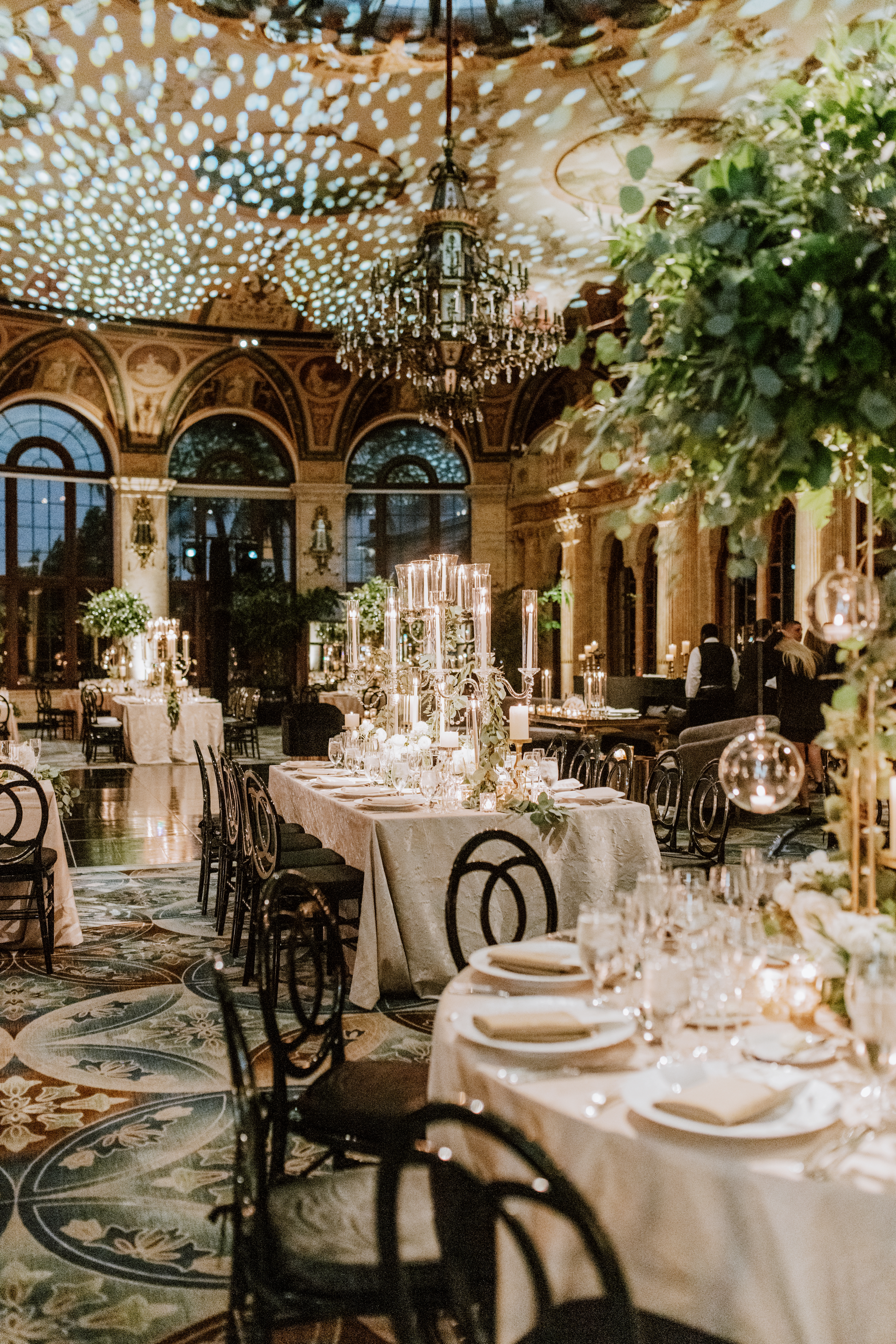 Elegant wedding reception in ornate ballroom with chandelier, long tables, greenery, and twinkling lights.