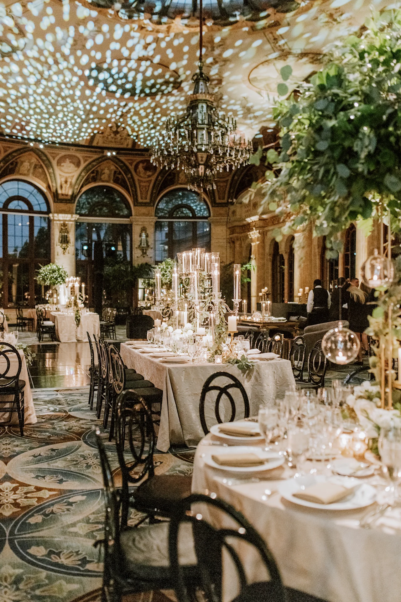 Elegant wedding reception in ornate ballroom with chandelier, long tables, greenery, and twinkling lights.