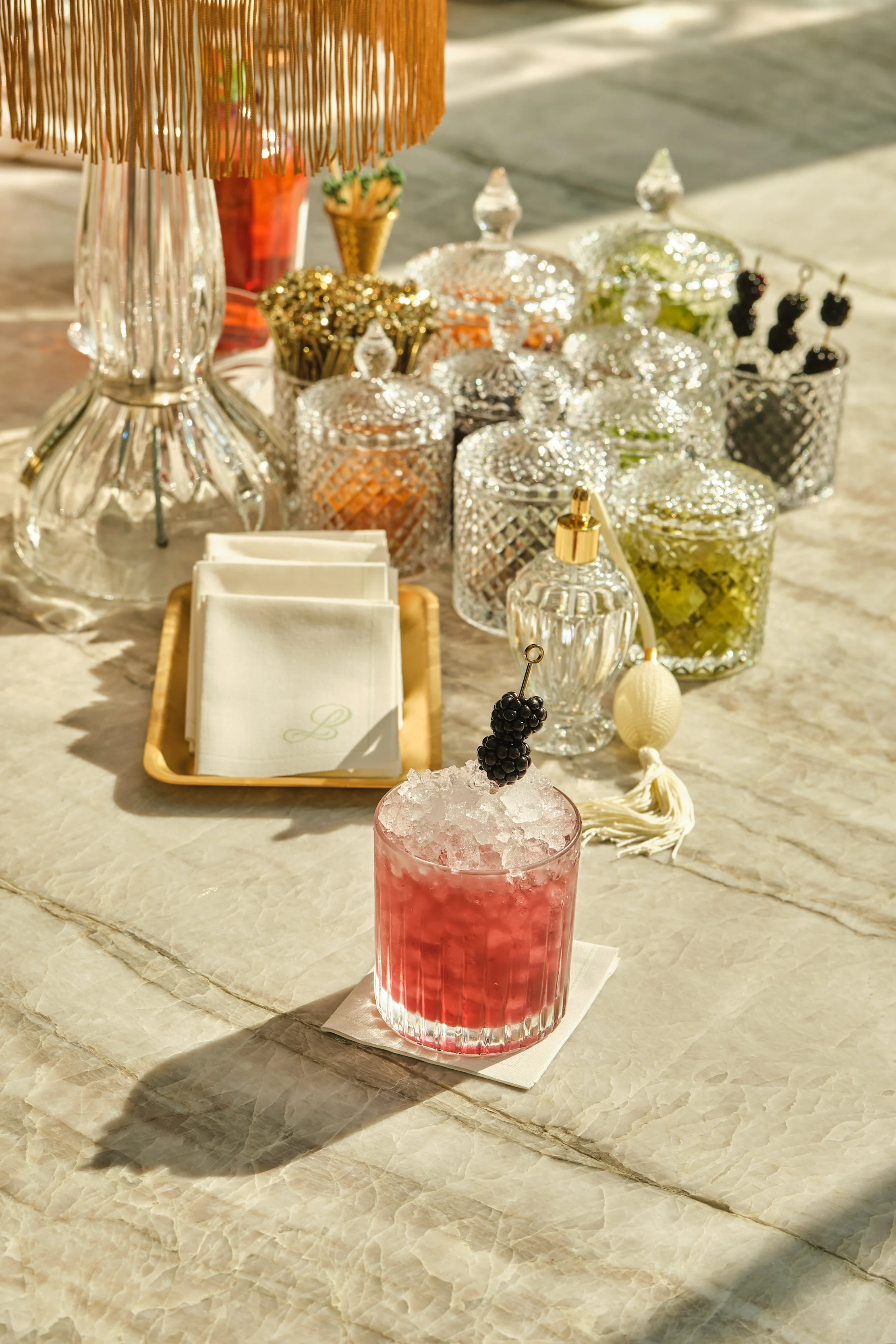 Red cocktail with crushed ice and blackberry garnish on marble table with crystal jars and lamp.