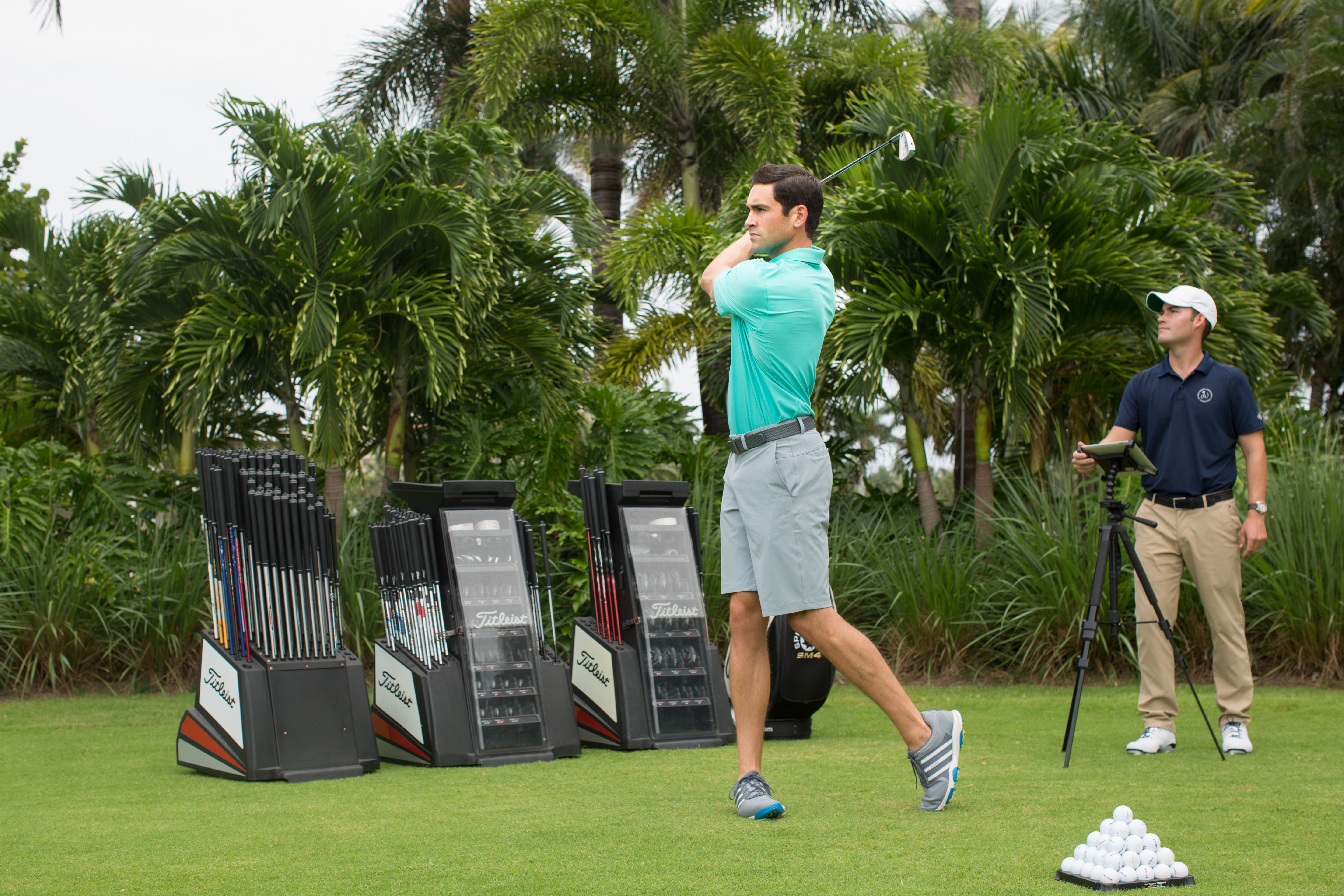 Golfer in turquoise shirt swinging club at driving range with palm trees and club displays.