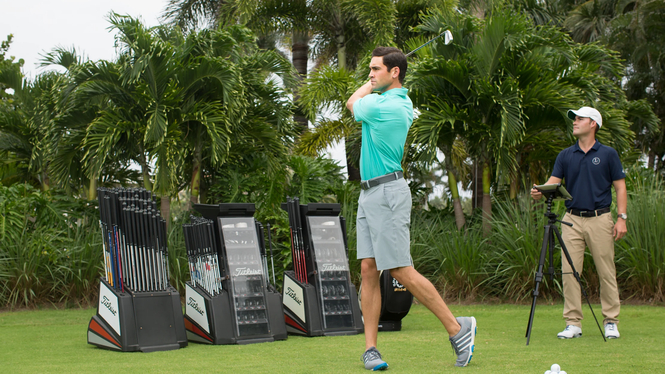 Golfer in turquoise shirt swinging club at driving range with palm trees and club displays.