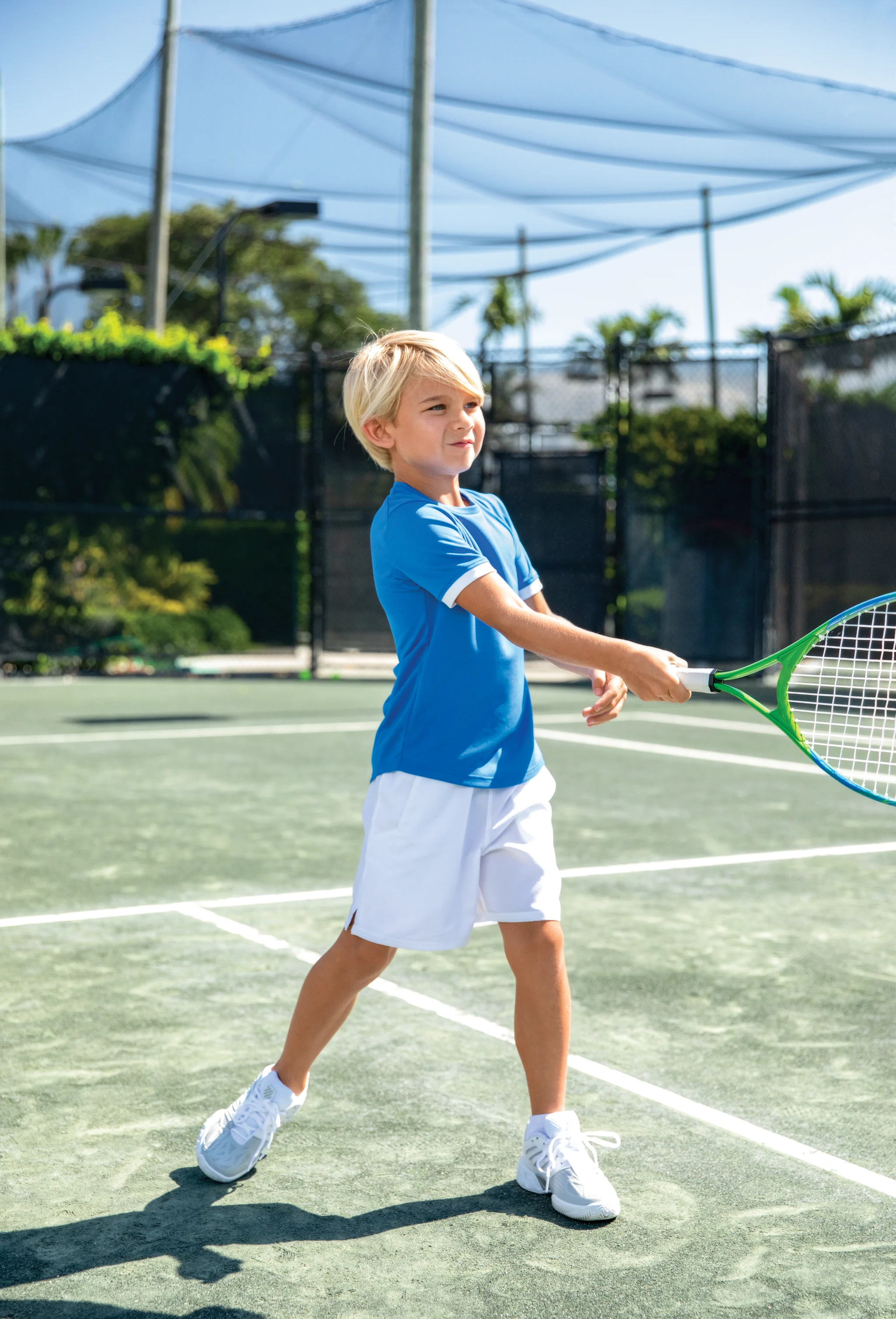 Young blonde boy in blue shirt and white shorts playing tennis on outdoor court with green racket.