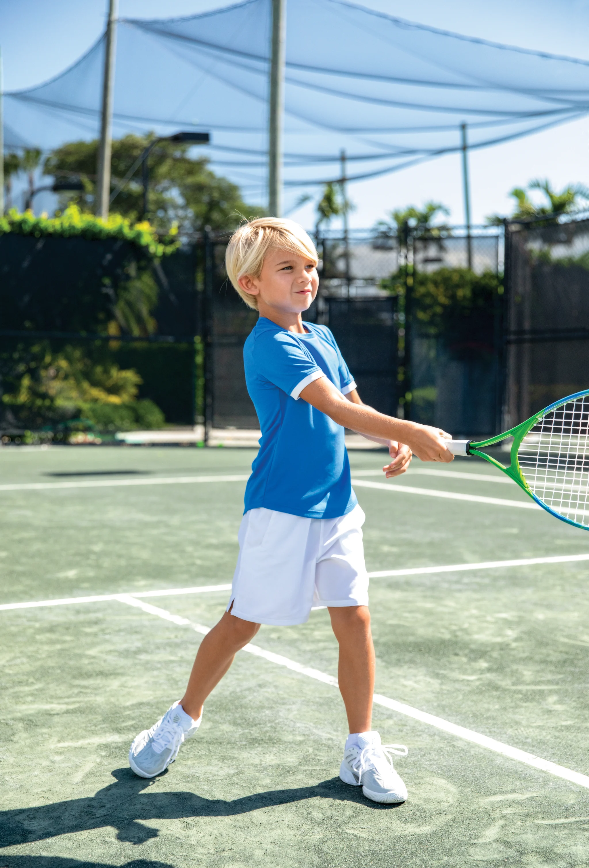 Young blonde boy in blue shirt and white shorts playing tennis on outdoor court with green racket.