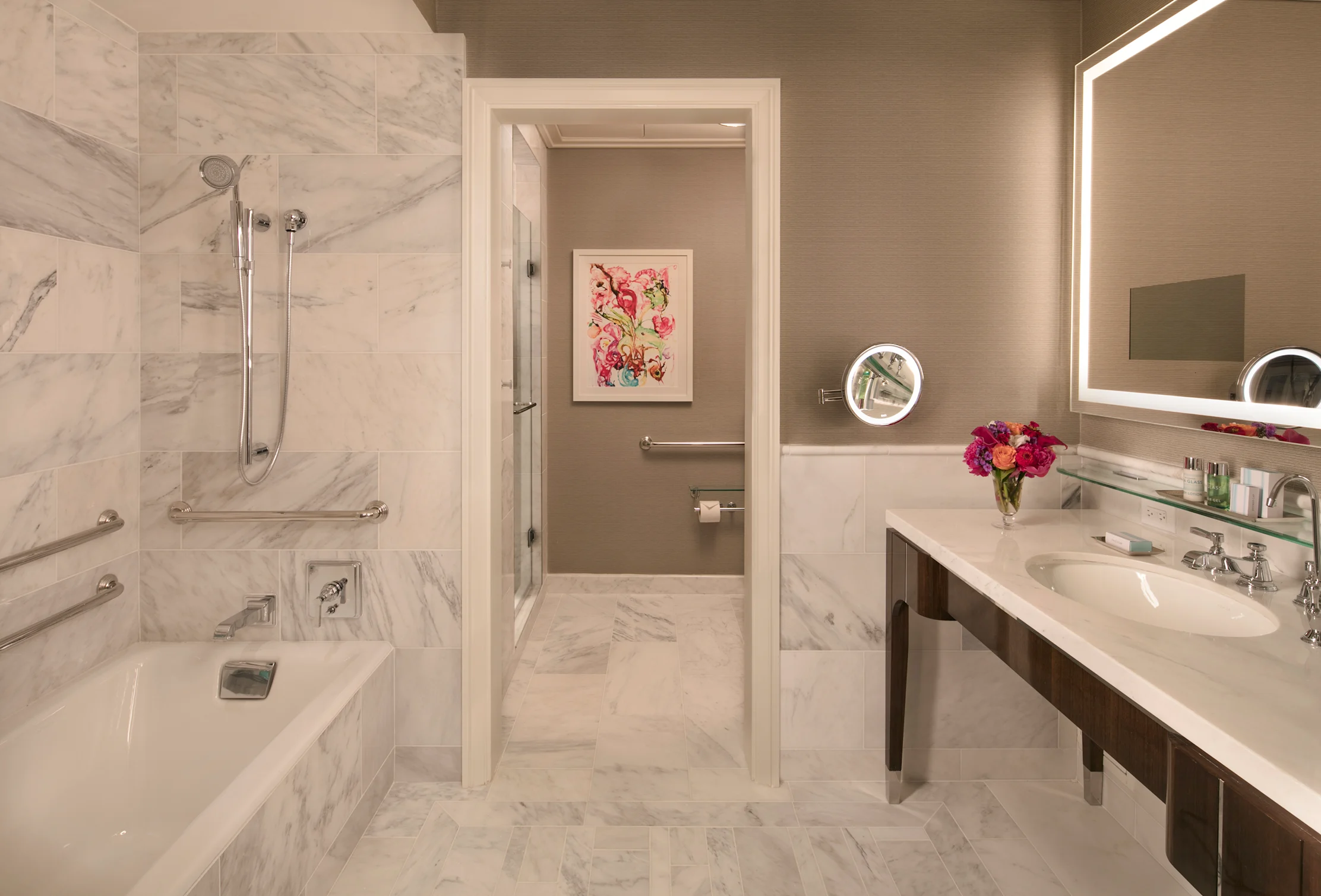 Elegant bathroom with white marble shower, beige walls, floral artwork, and modern vanity with flowers.
