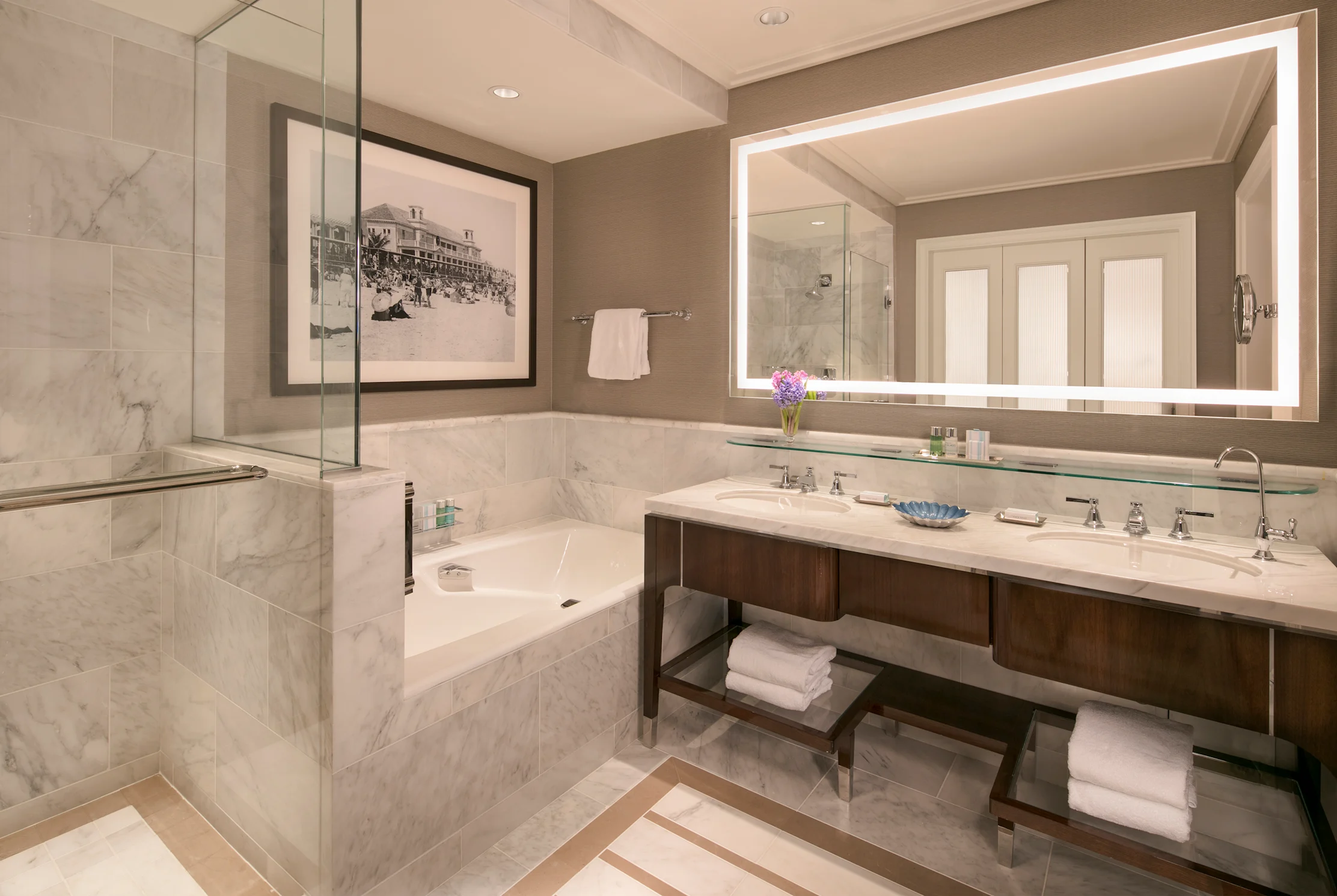 Luxury hotel bathroom with marble tub, double vanity, large backlit mirror, and glass shower.