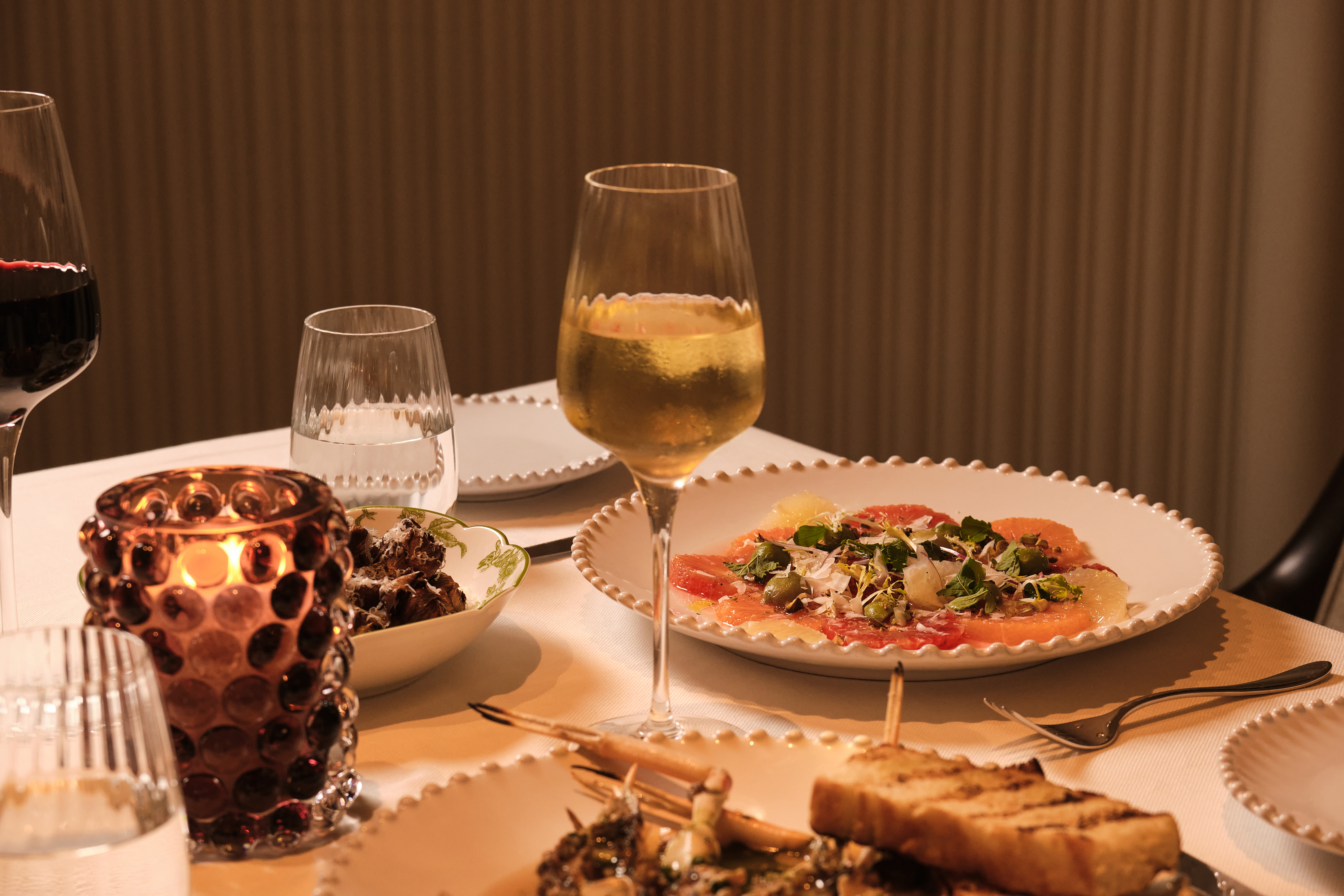Elegant dinner table with wine glasses, citrus carpaccio, and candlelight against brown backdrop
