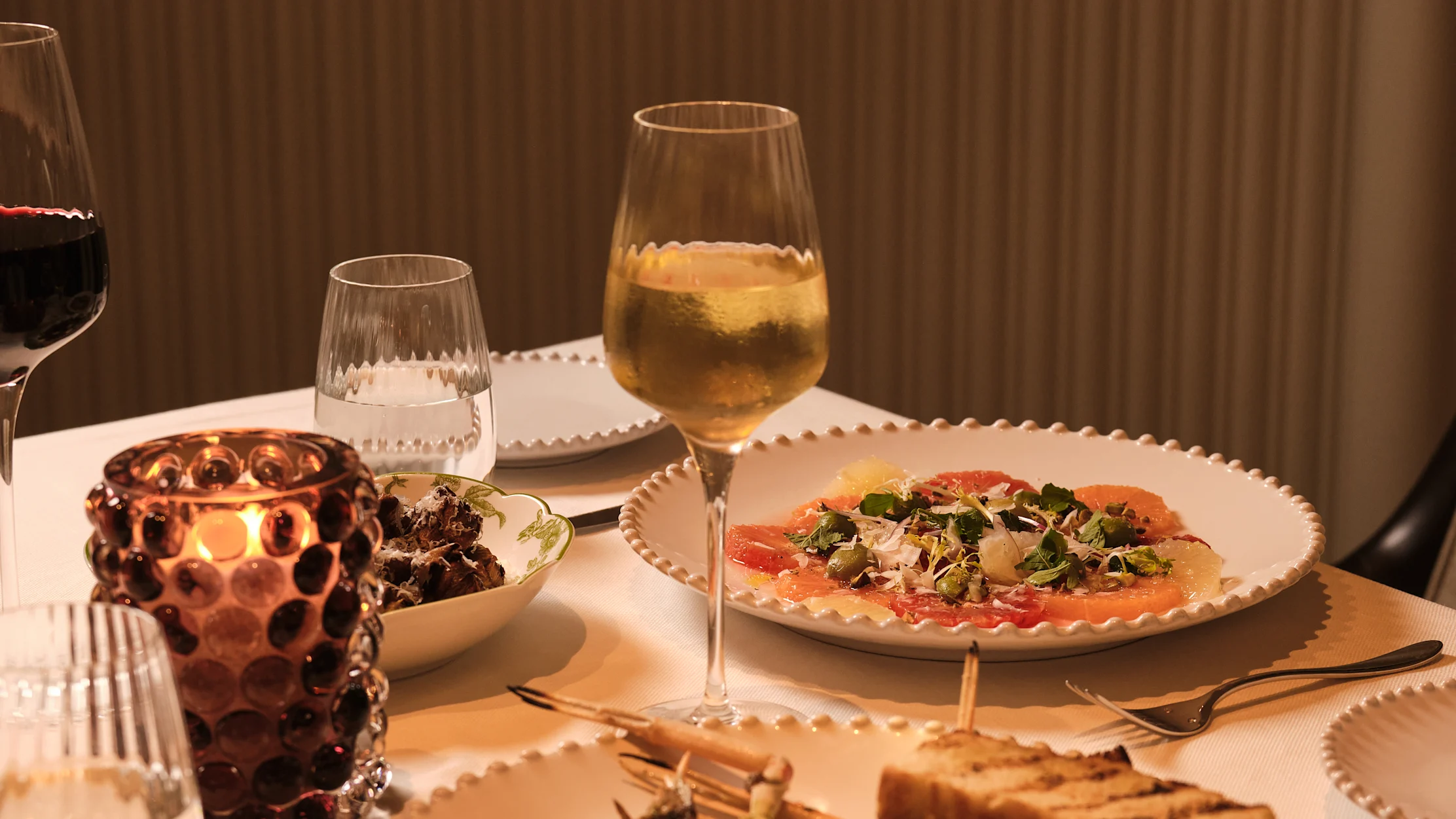 Elegant dinner table with wine glasses, citrus carpaccio, and candlelight against brown backdrop