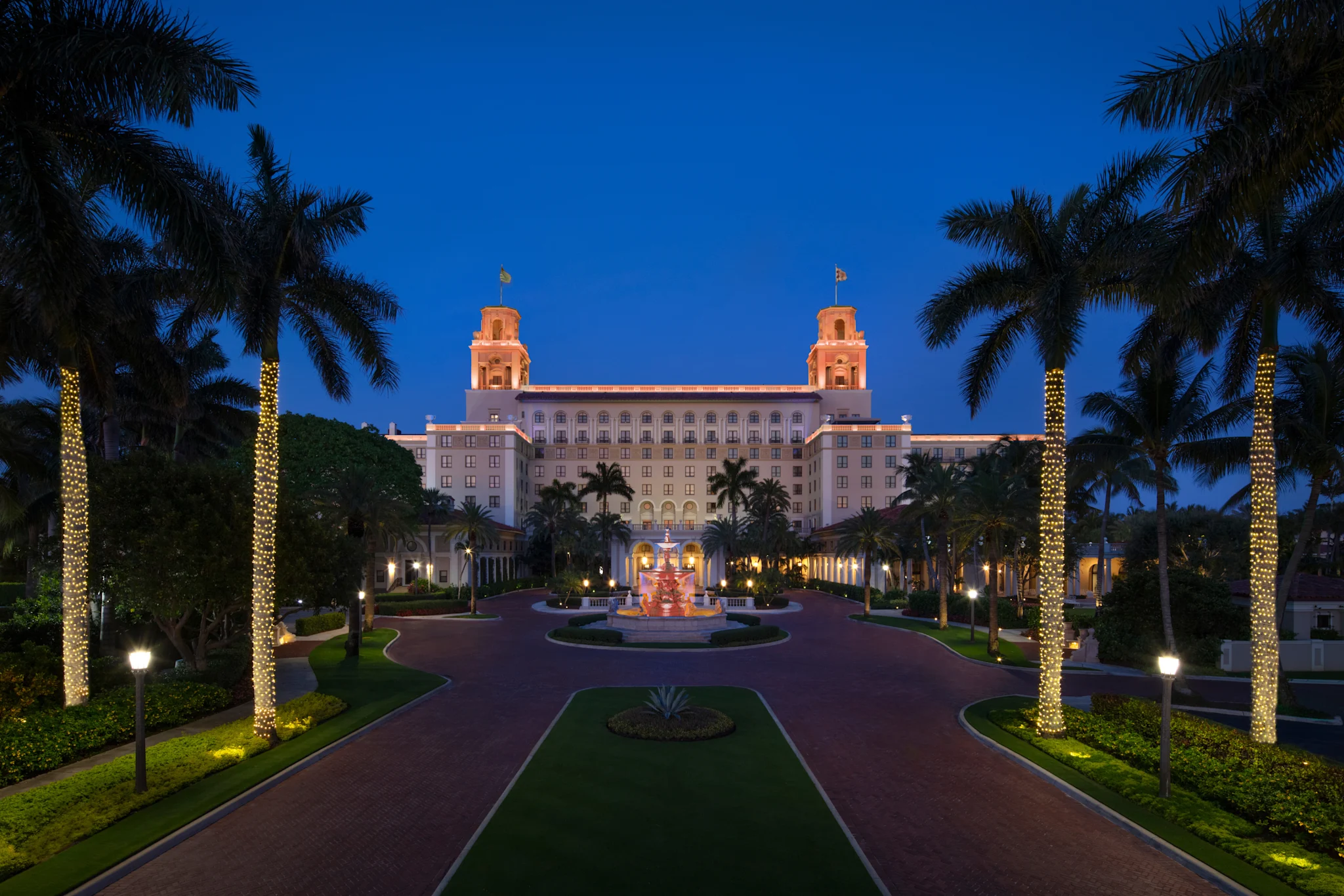 Illuminated hotel The Breakers Palm Beach with twin towers at dusk, framed by palm trees with string lights and fountain.
