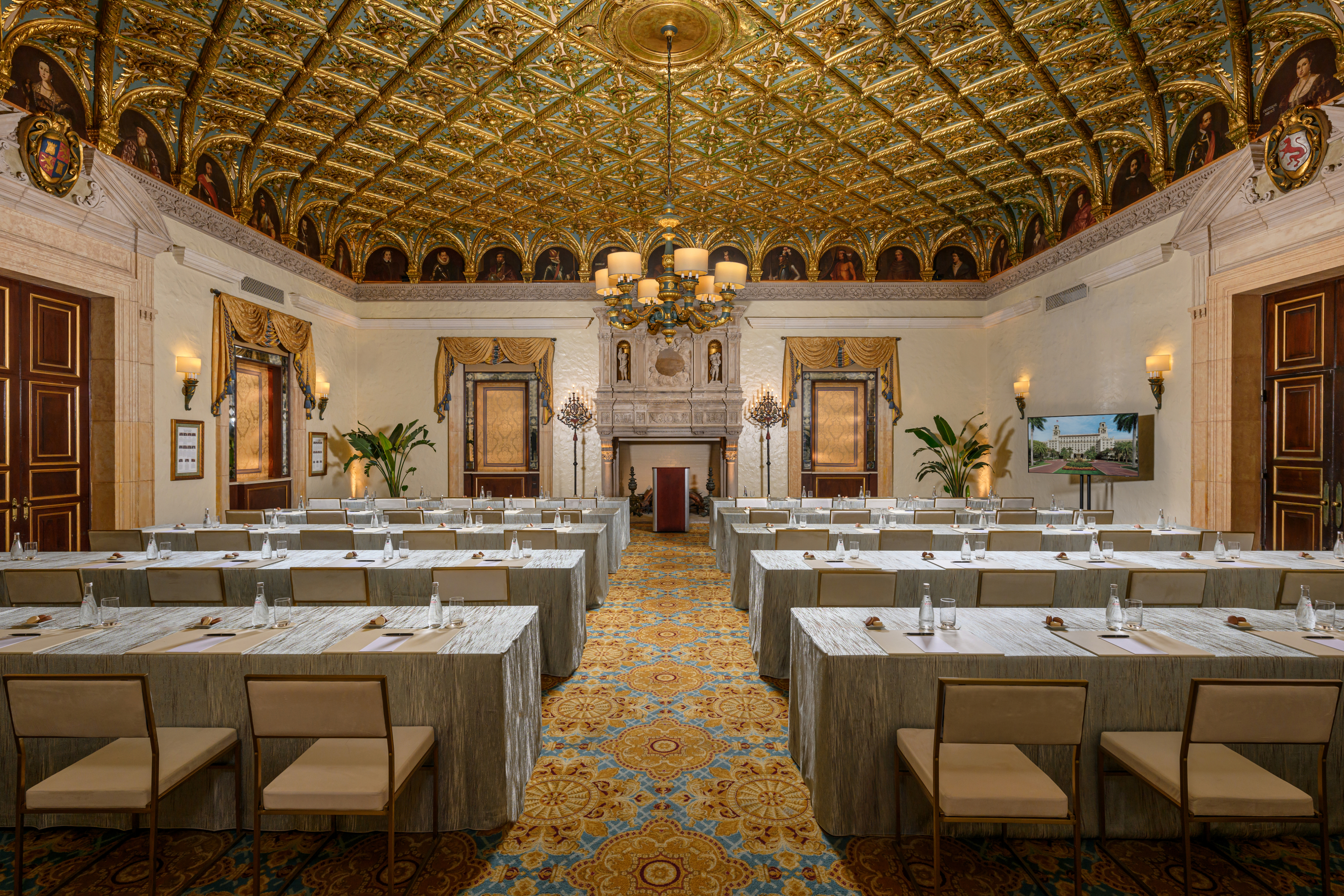 Ornate ballroom with gold-patterned ceiling, chandelier, and rows of tables set for a meeting.
