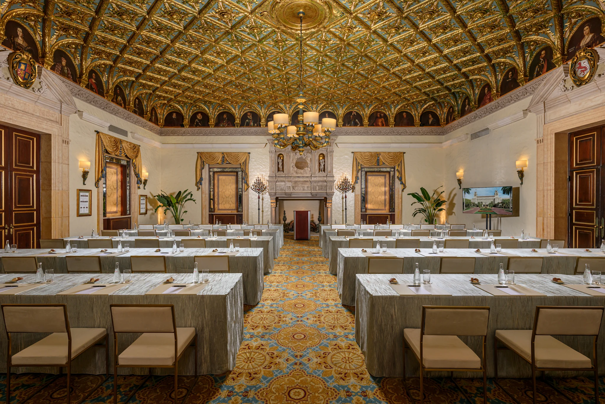 Ornate ballroom with gold-patterned ceiling, chandelier, and rows of tables set for a meeting.