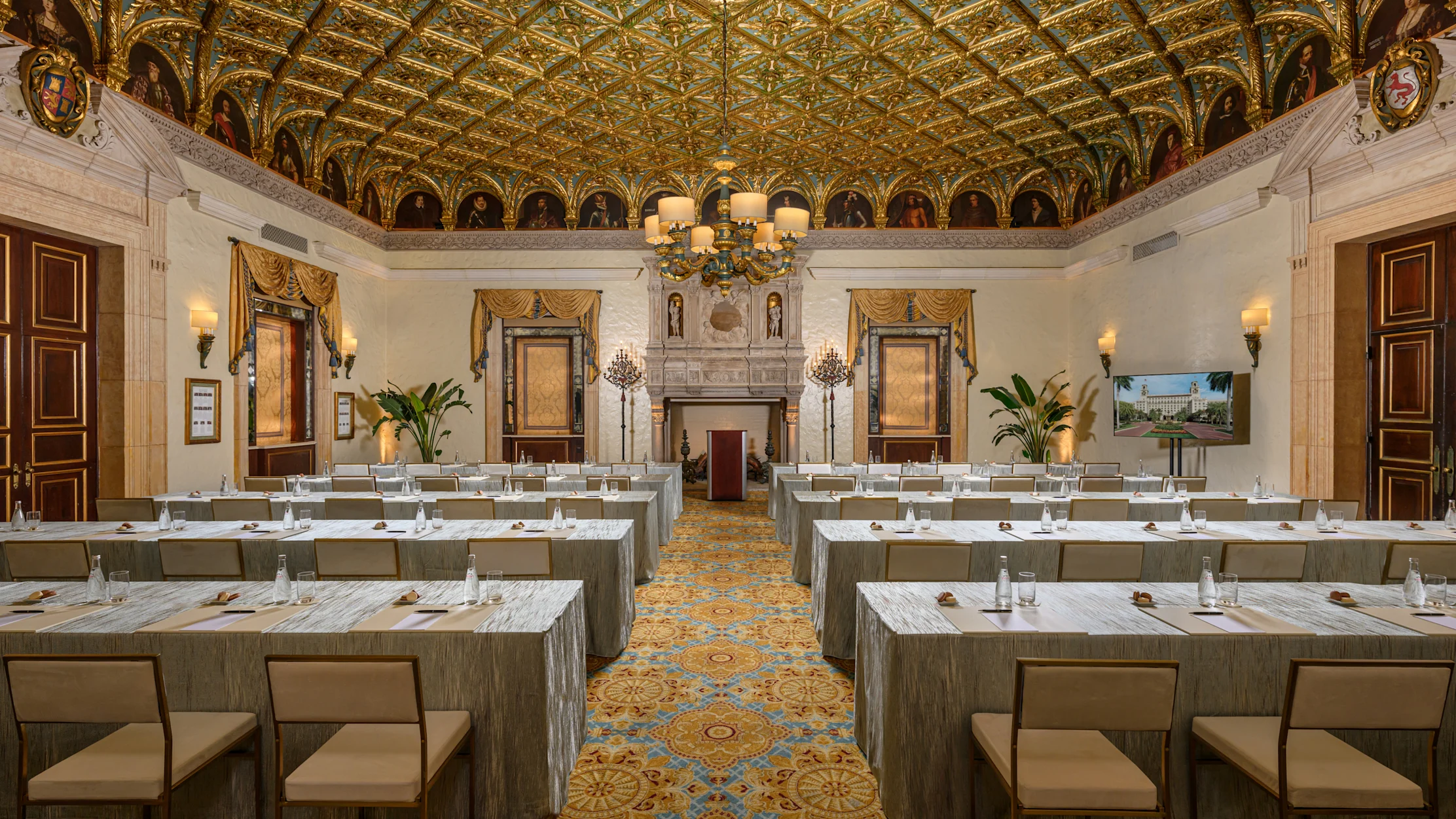 Ornate ballroom with gold-patterned ceiling, chandelier, and rows of tables set for a meeting.