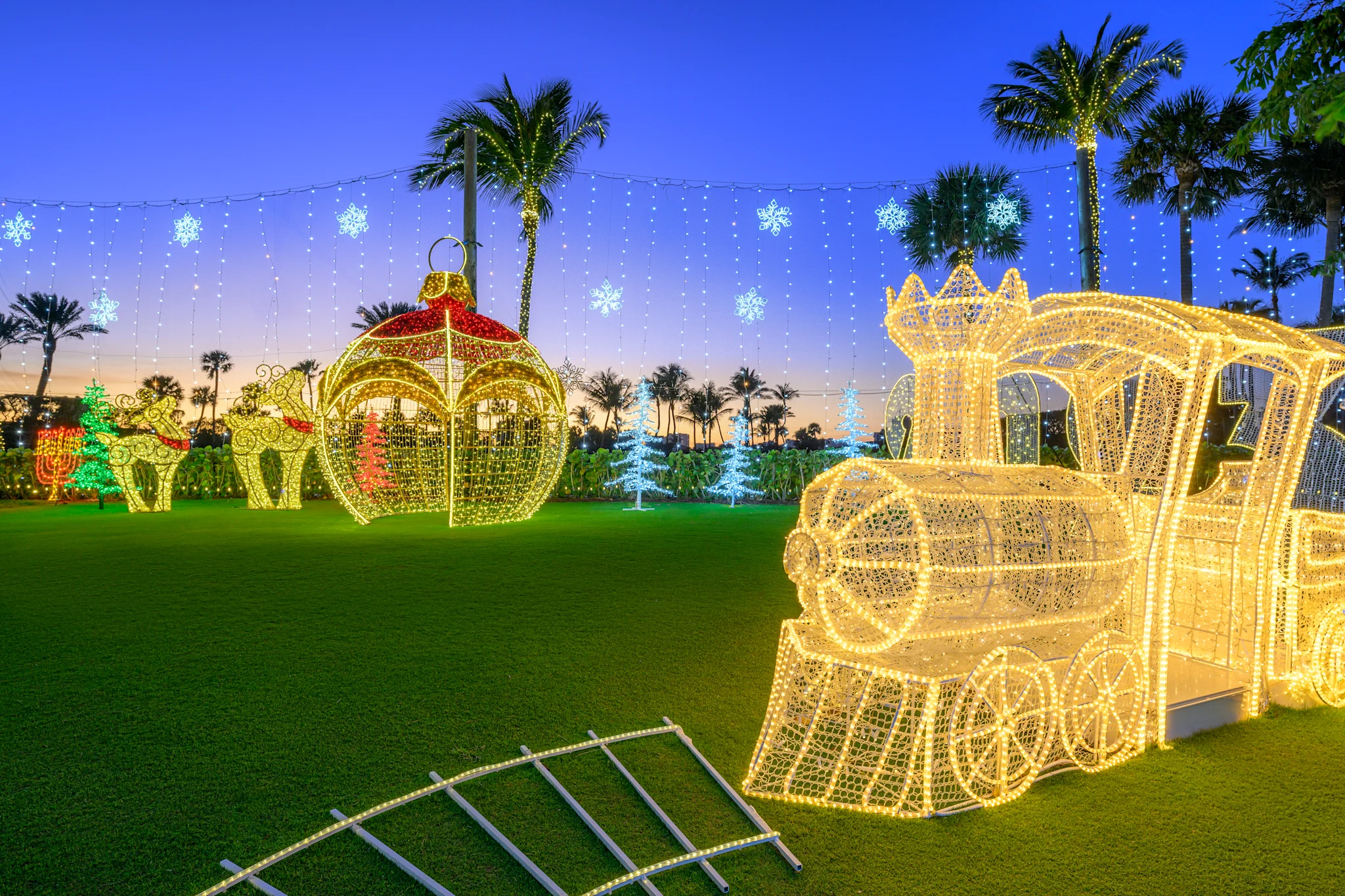 Illuminated Christmas decorations including a golden train and ornament on lawn with palm trees at dusk.