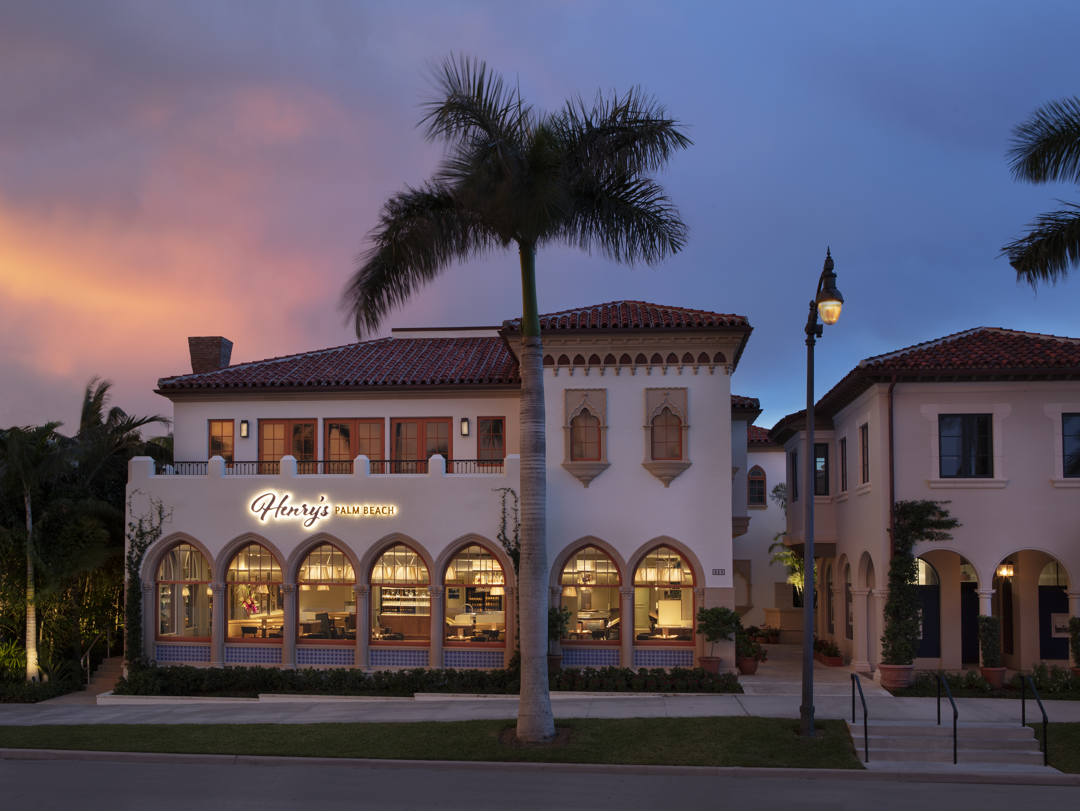 Mediterranean-style building with arched windows and red tile roof at dusk with palm tree.