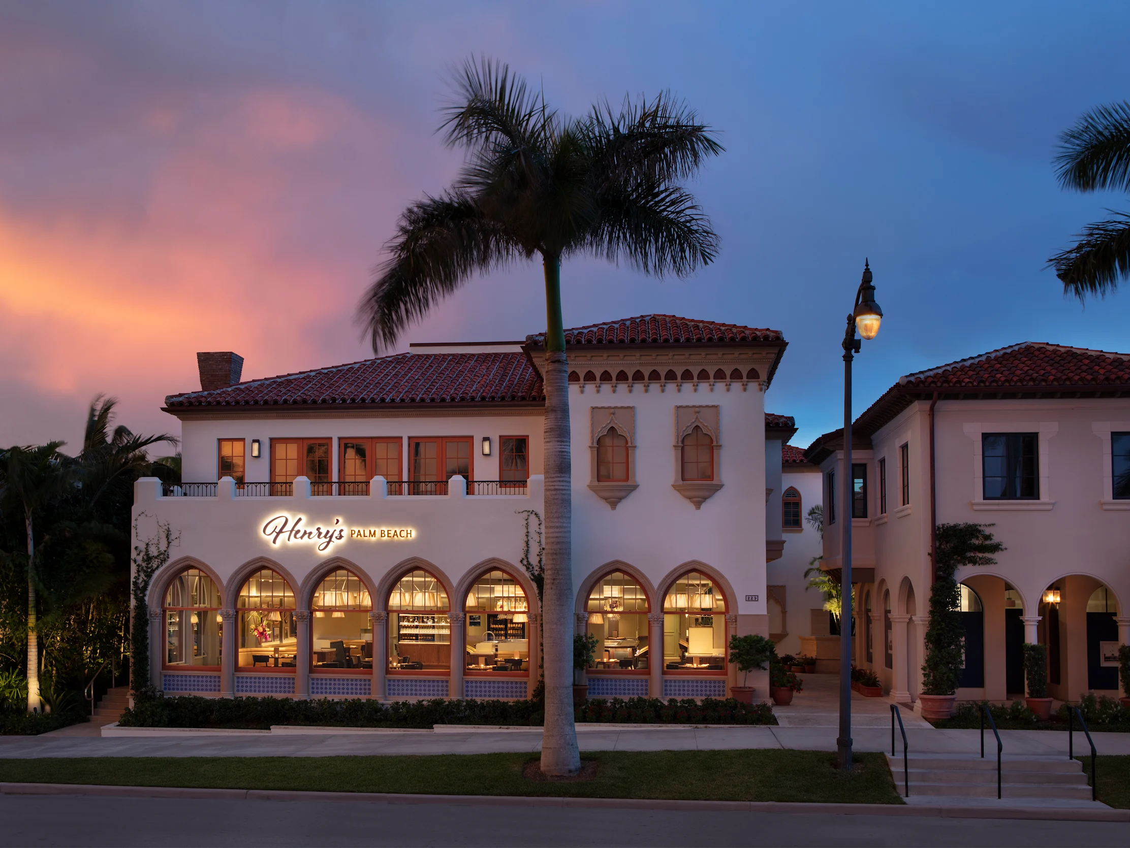 Mediterranean-style building with arched windows and red tile roof at dusk with palm tree.
