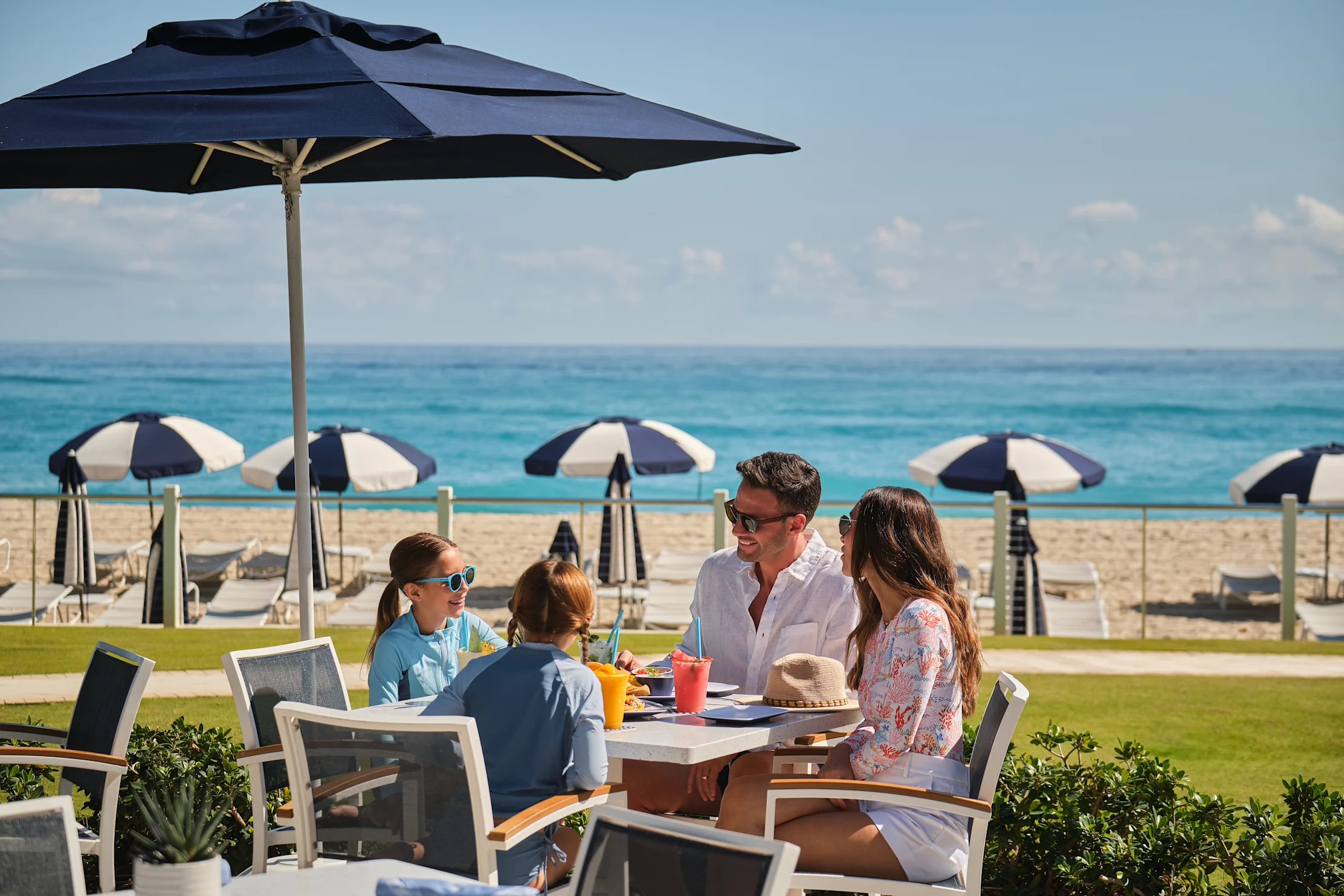 Family dining at beachfront table under blue umbrella with ocean and striped beach umbrellas in background.