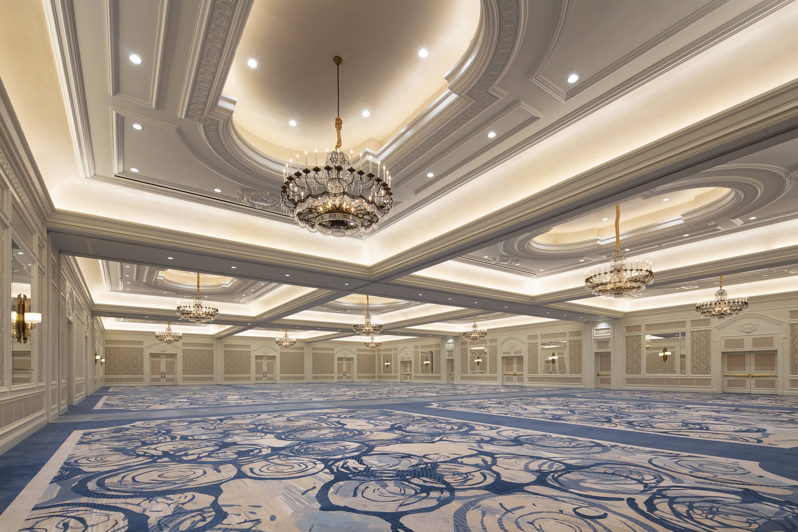 Elegant ballroom with ornate ceiling, crystal chandeliers, and blue patterned carpet in neutral tones.