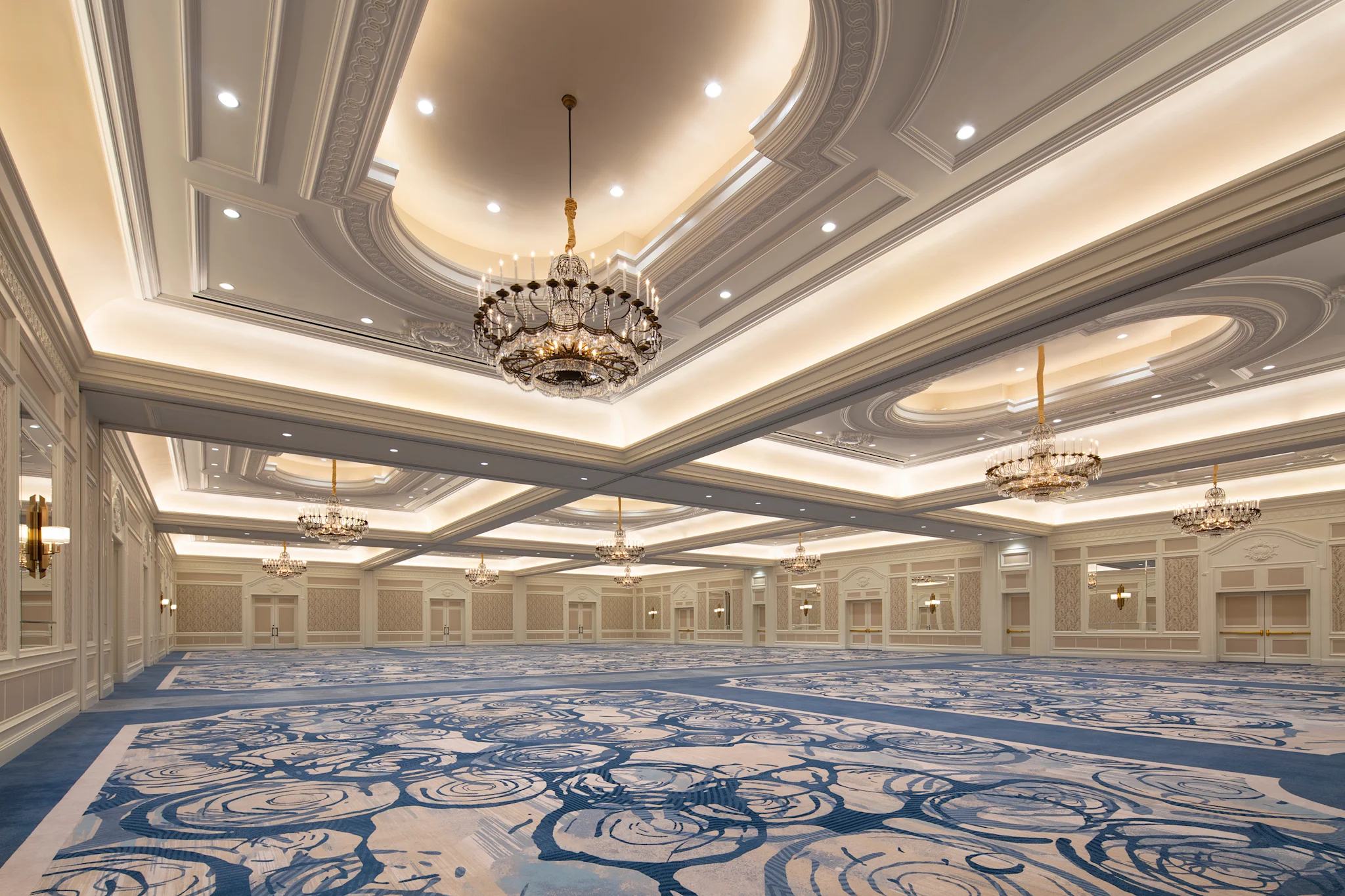 Elegant ballroom with ornate ceiling, crystal chandeliers, and blue patterned carpet in neutral tones.