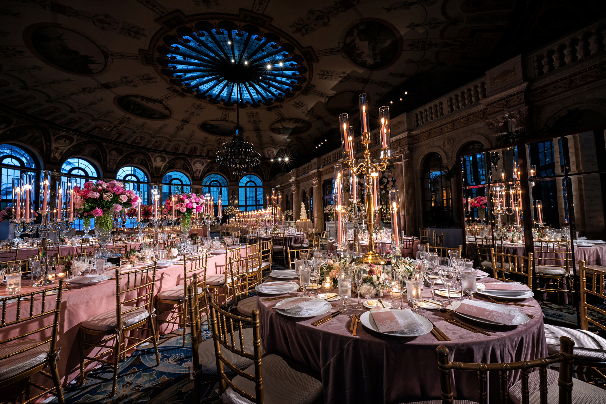 Elegant ballroom with ornate ceiling, blue chandelier, pink linens, gold chairs, and tall candelabras.