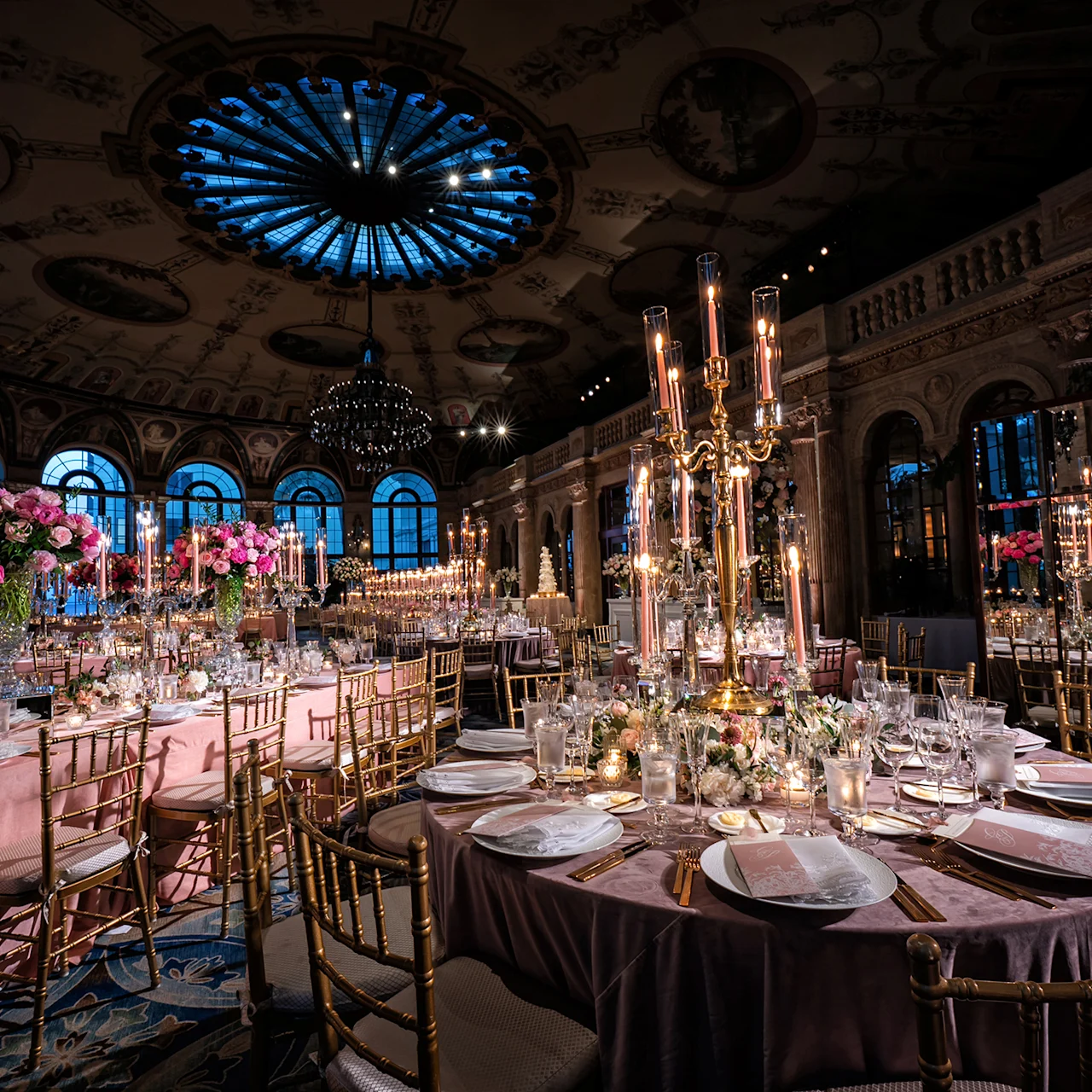 Elegant ballroom with ornate ceiling, blue chandelier, pink linens, gold chairs, and tall candelabras.