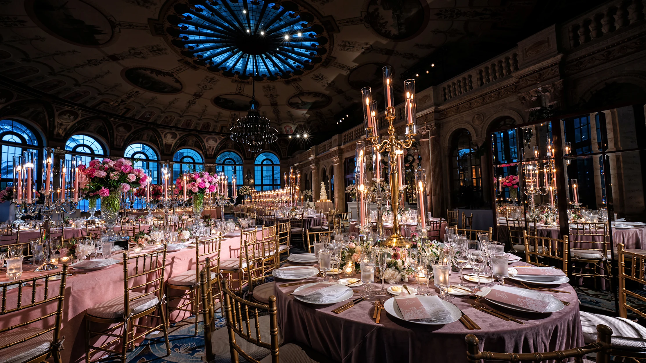Elegant ballroom with ornate ceiling, blue chandelier, pink linens, gold chairs, and tall candelabras.