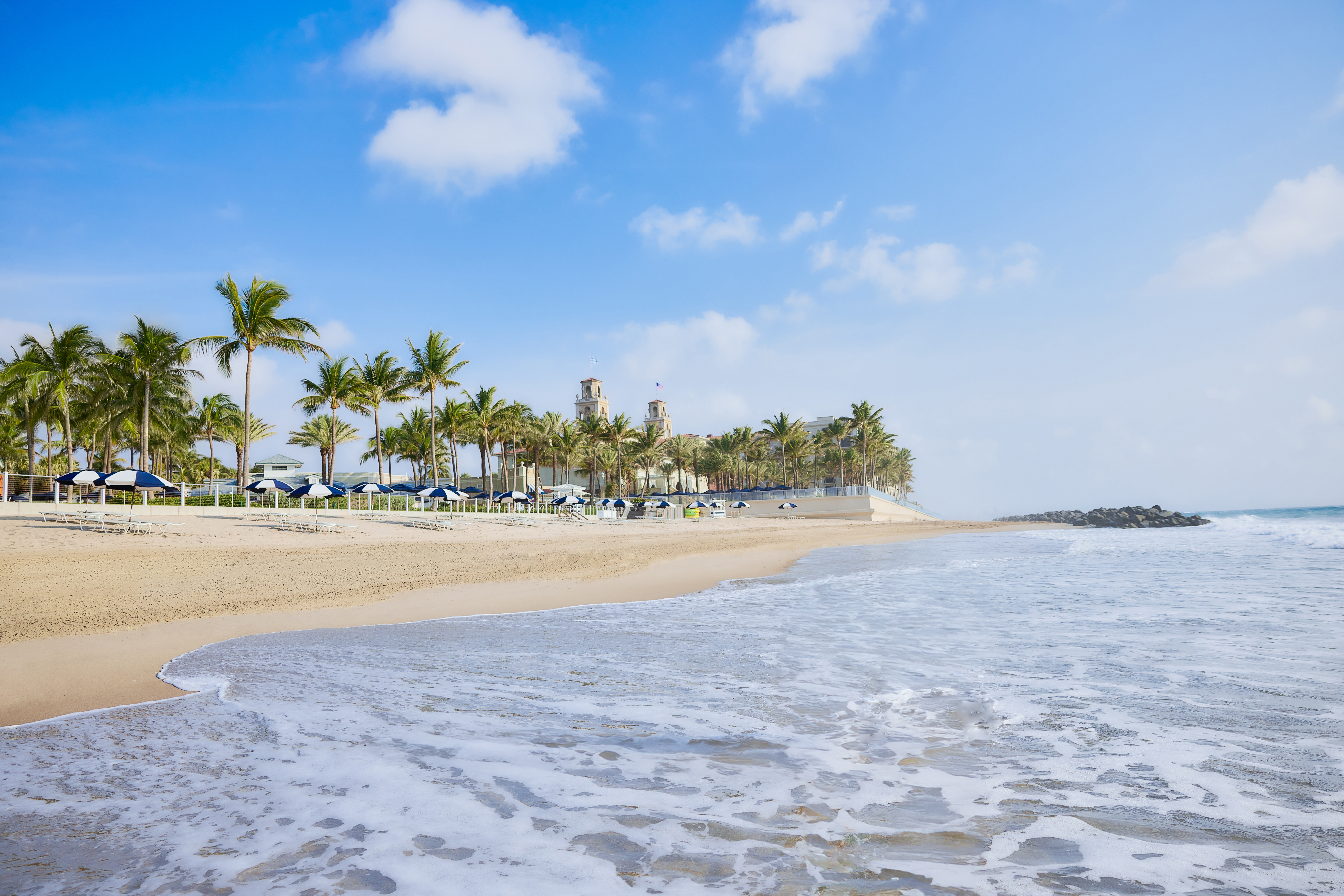 Tropical beach with palm trees, blue umbrellas, and gentle waves under a partly cloudy sky.
