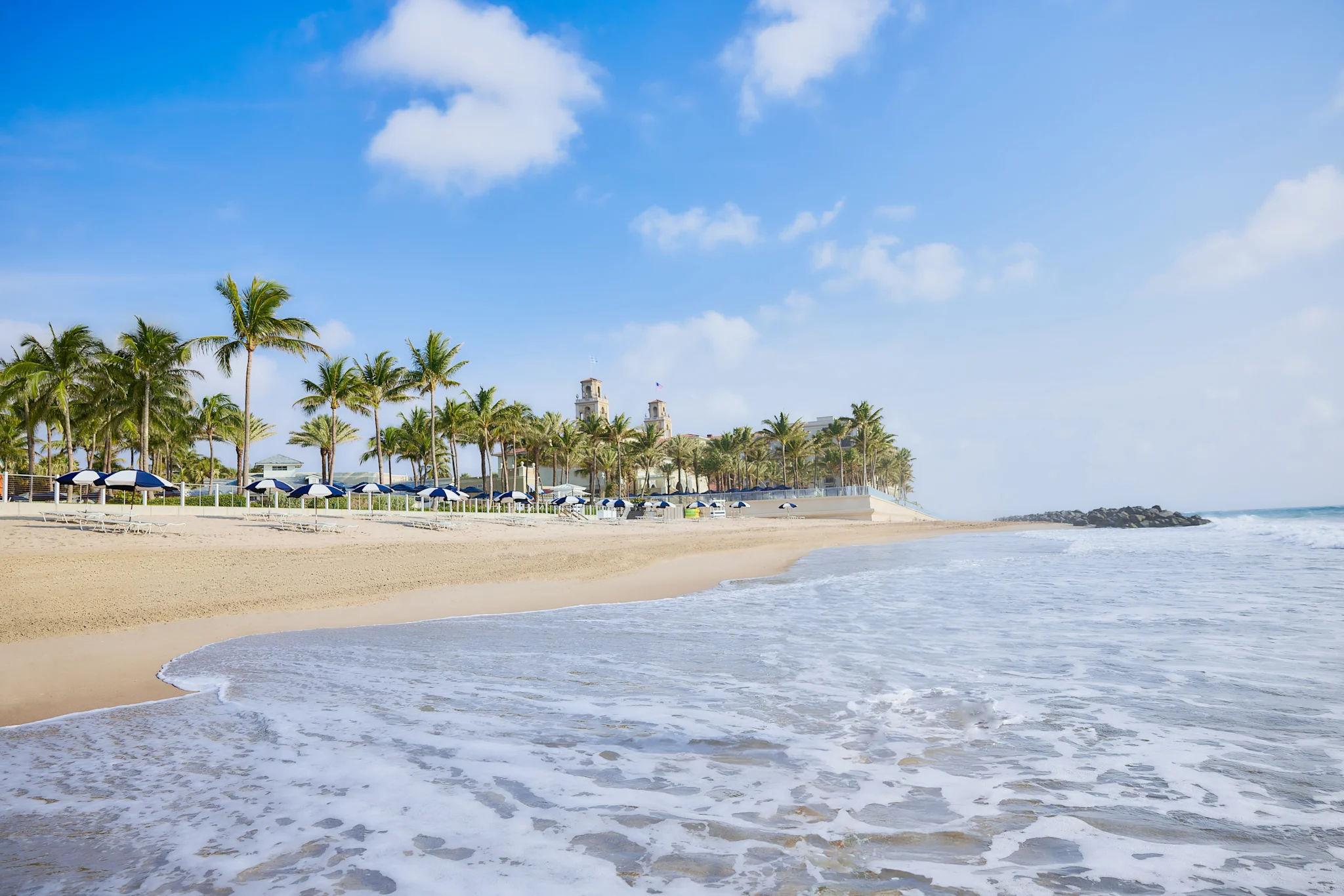 Tropical beach with palm trees, blue umbrellas, and gentle waves under a partly cloudy sky.