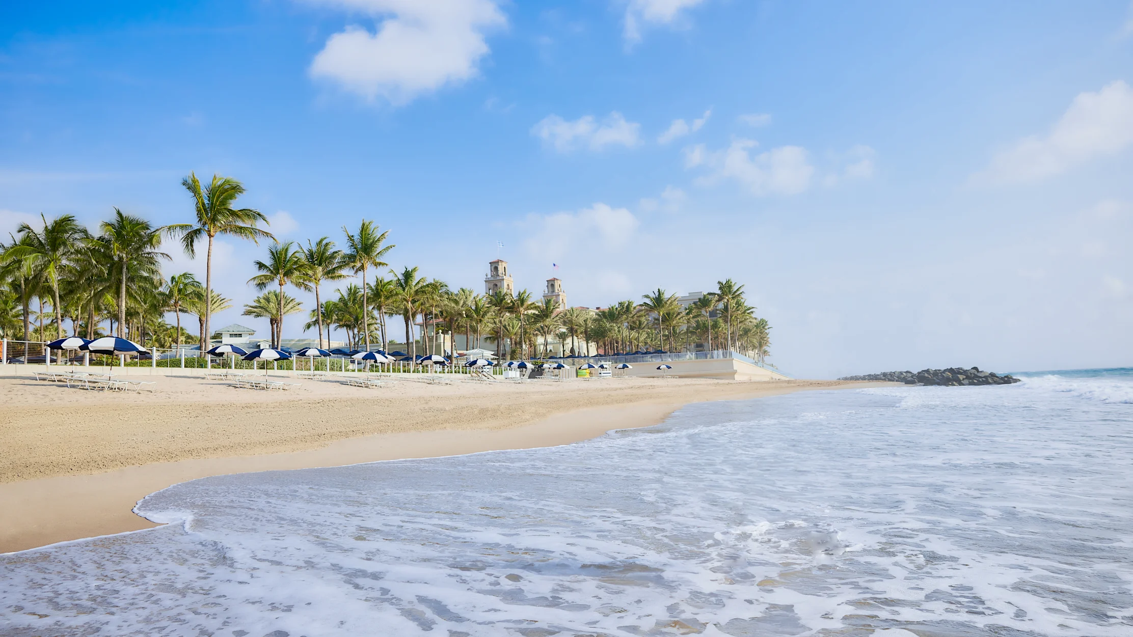 Tropical beach with palm trees, blue umbrellas, and gentle waves under a partly cloudy sky.