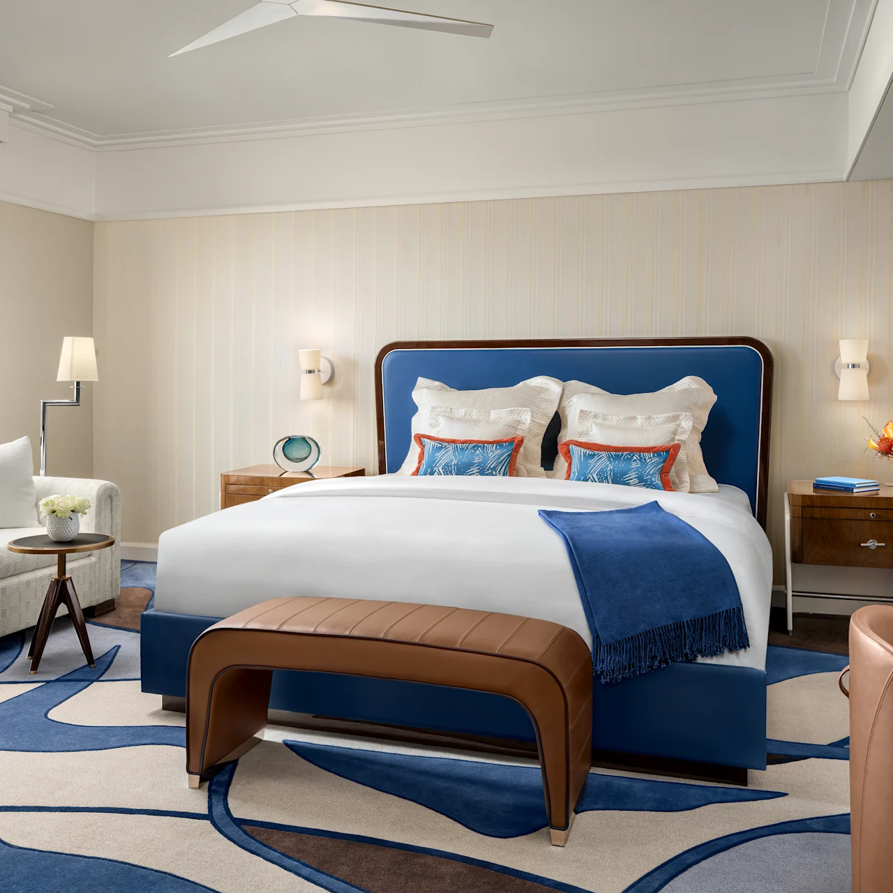 Elegant hotel bedroom with blue upholstered bed, white linens, modern furniture, and city views.
