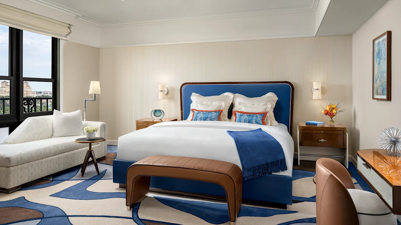 Elegant hotel bedroom with blue upholstered bed, white linens, modern furniture, and city views.