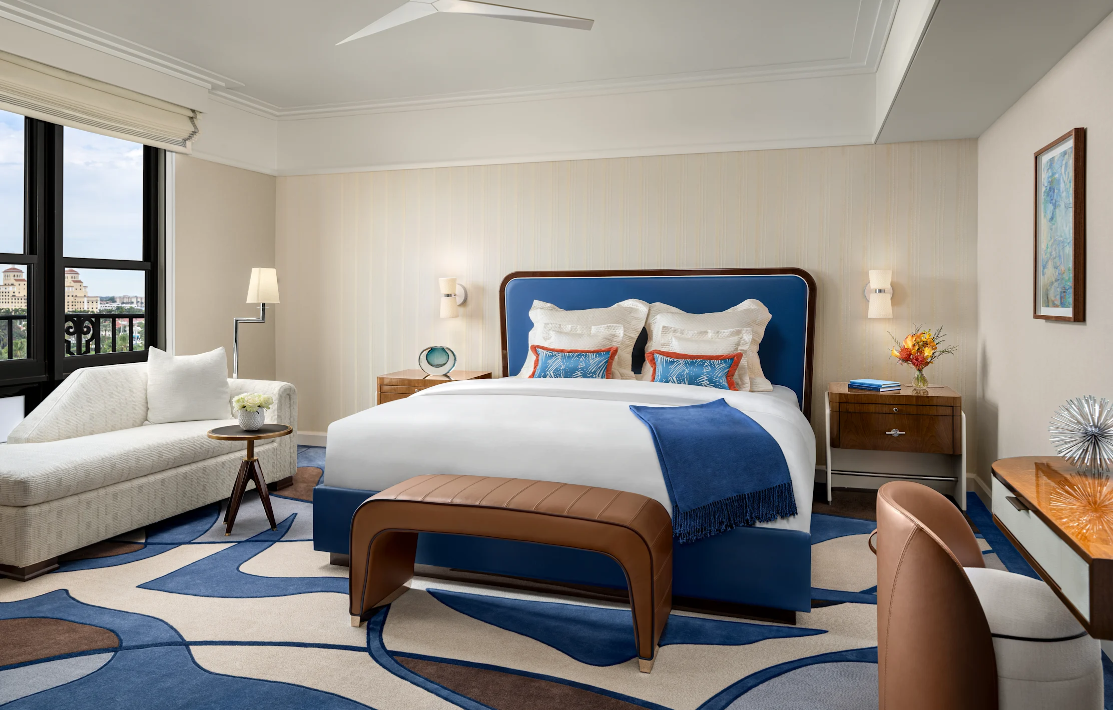 Elegant hotel bedroom with blue upholstered bed, white linens, modern furniture, and city views.