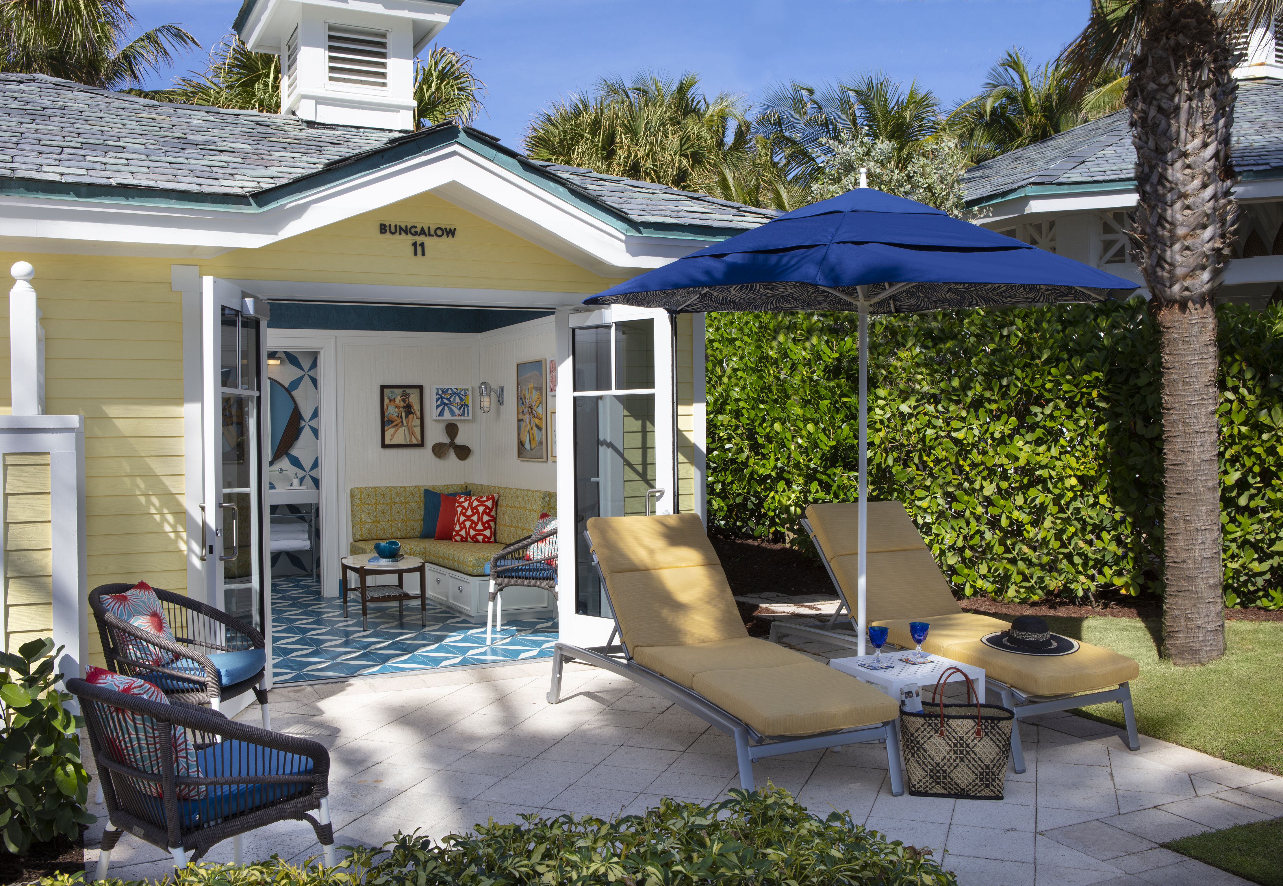 Yellow bungalow with open doors revealing interior, patio with lounge chairs under blue umbrella, palm trees.