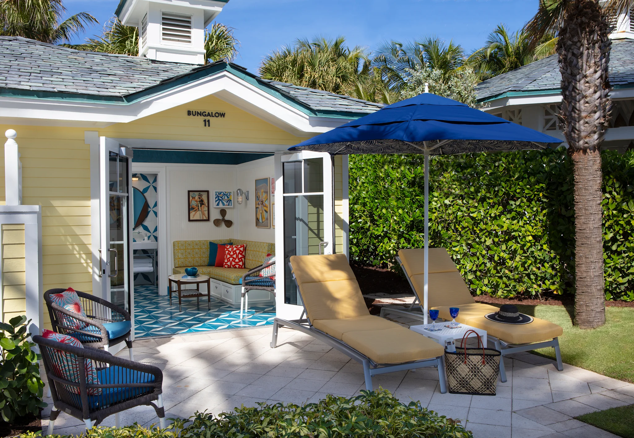 Yellow bungalow with open doors revealing interior, patio with lounge chairs under blue umbrella, palm trees.