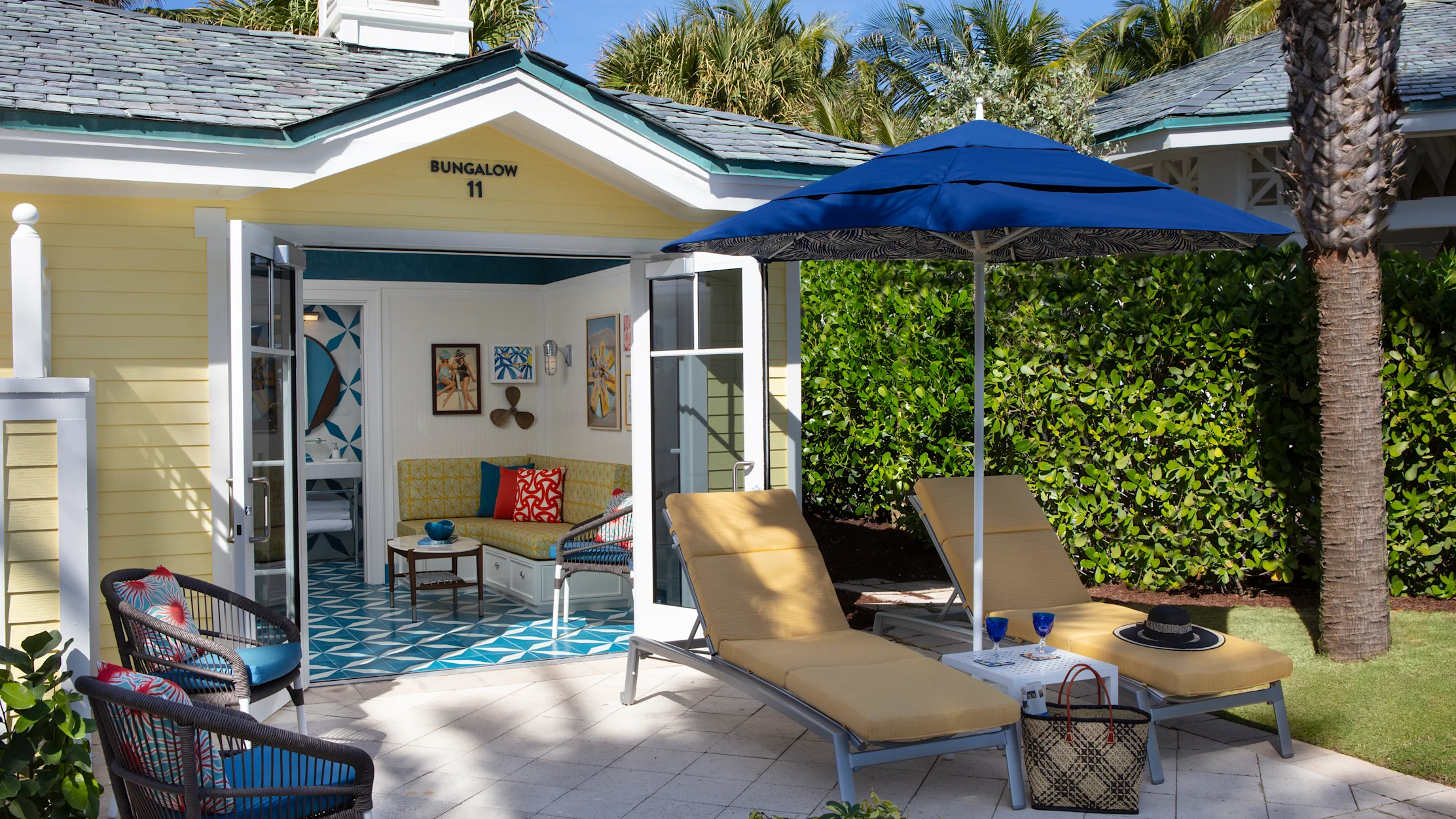 Yellow bungalow with open doors revealing interior, patio with lounge chairs under blue umbrella, palm trees.
