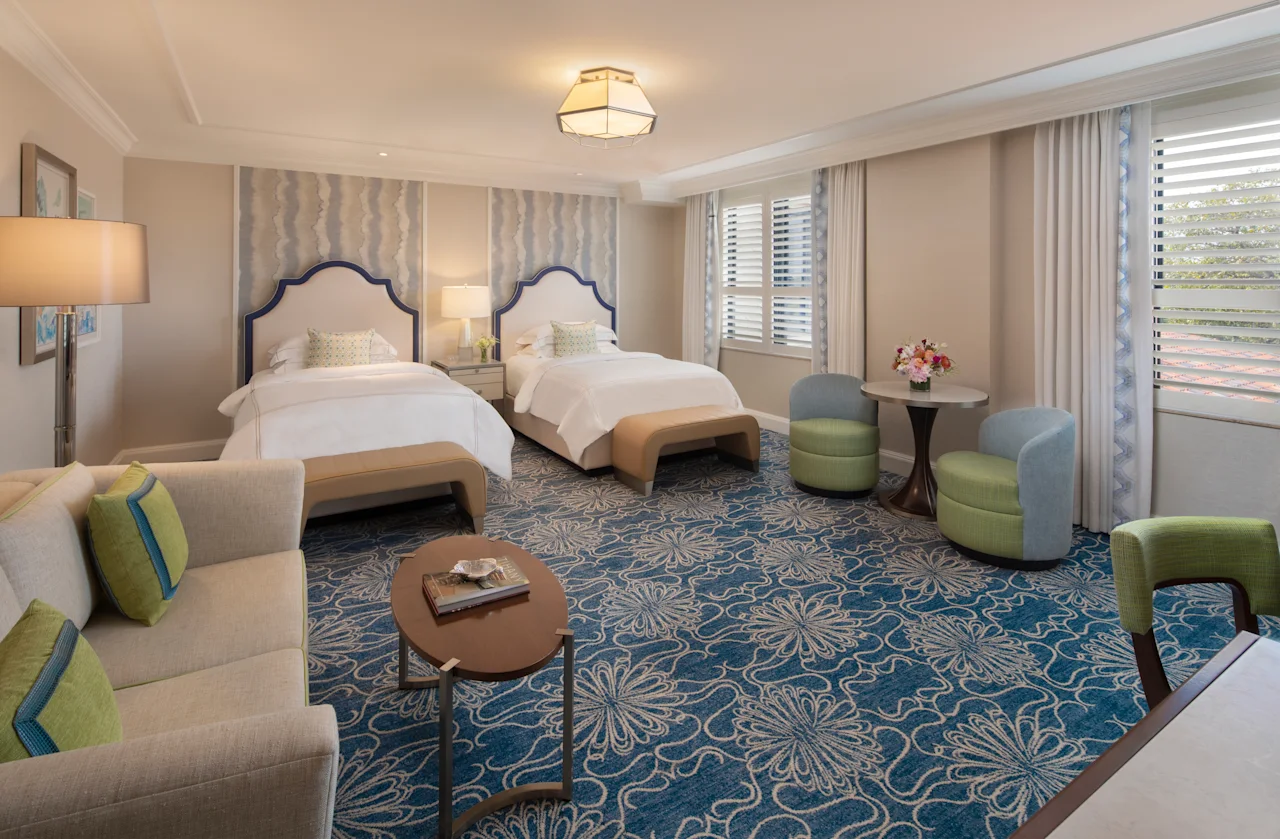 Elegant hotel room with two beds, blue patterned carpet, beige seating, and green accent chairs.