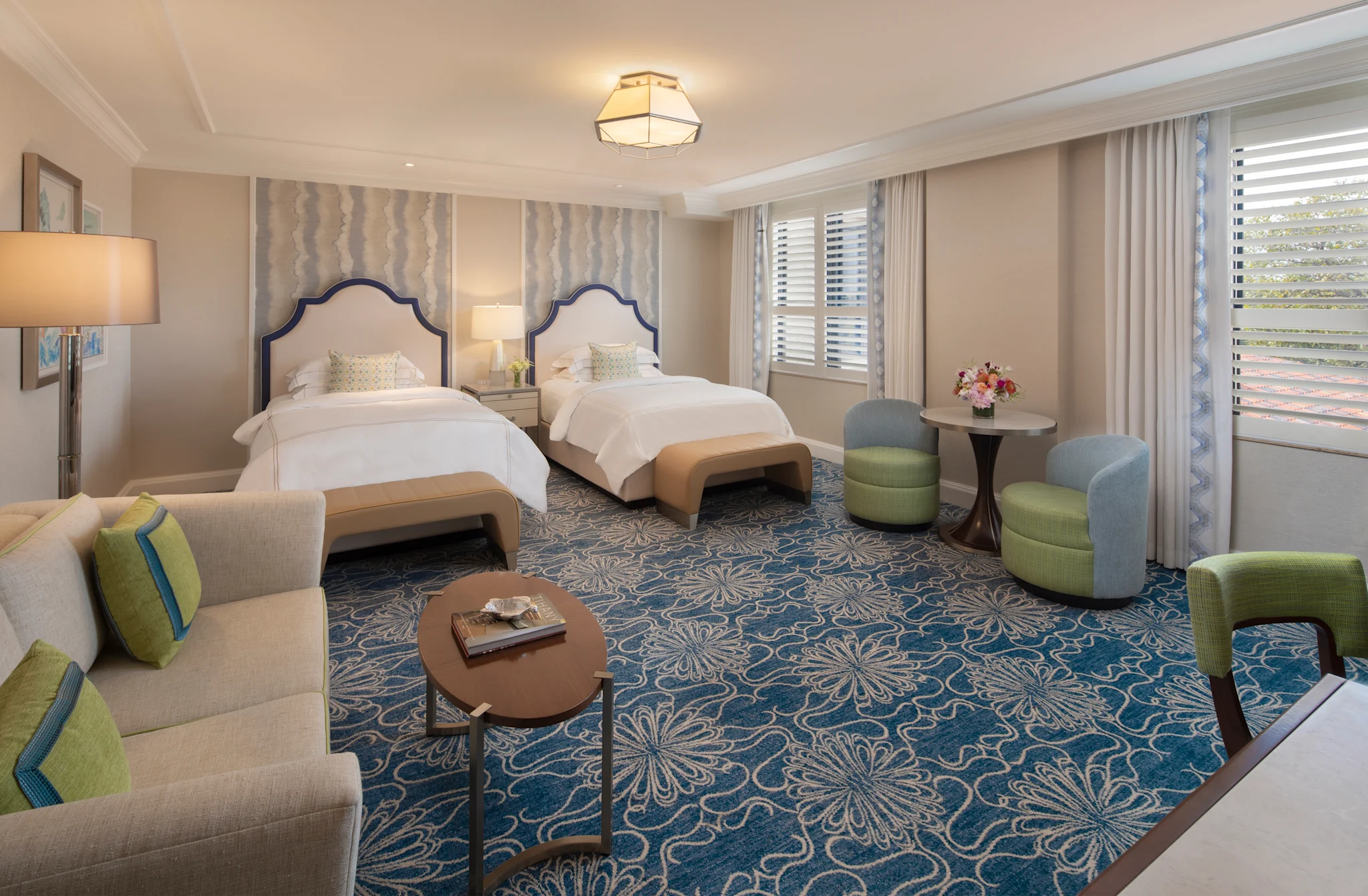 Elegant hotel room with two beds, blue patterned carpet, beige seating, and green accent chairs.