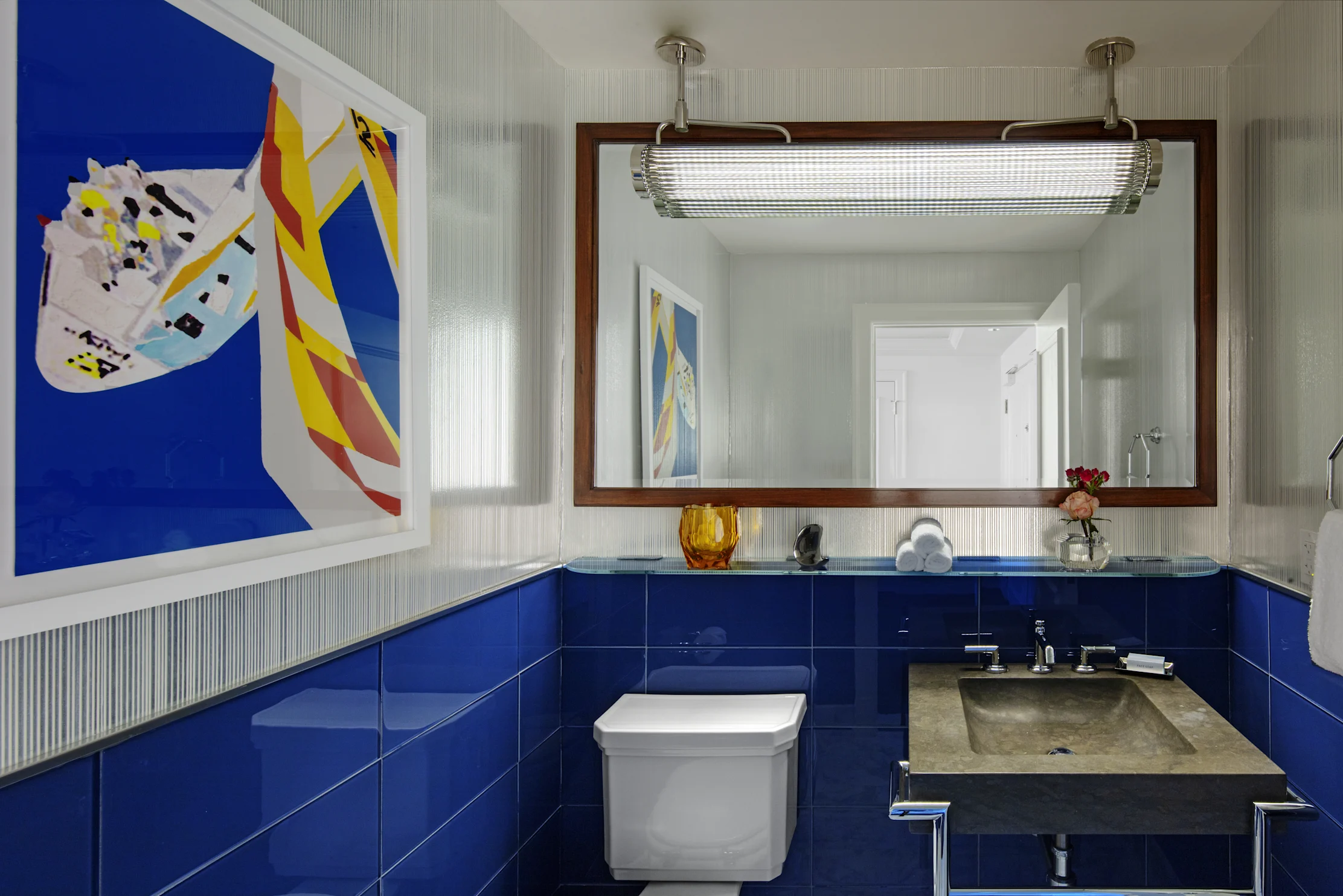 Modern bathroom with blue tile walls, abstract artwork, wood-framed mirror, and chrome light fixture.
