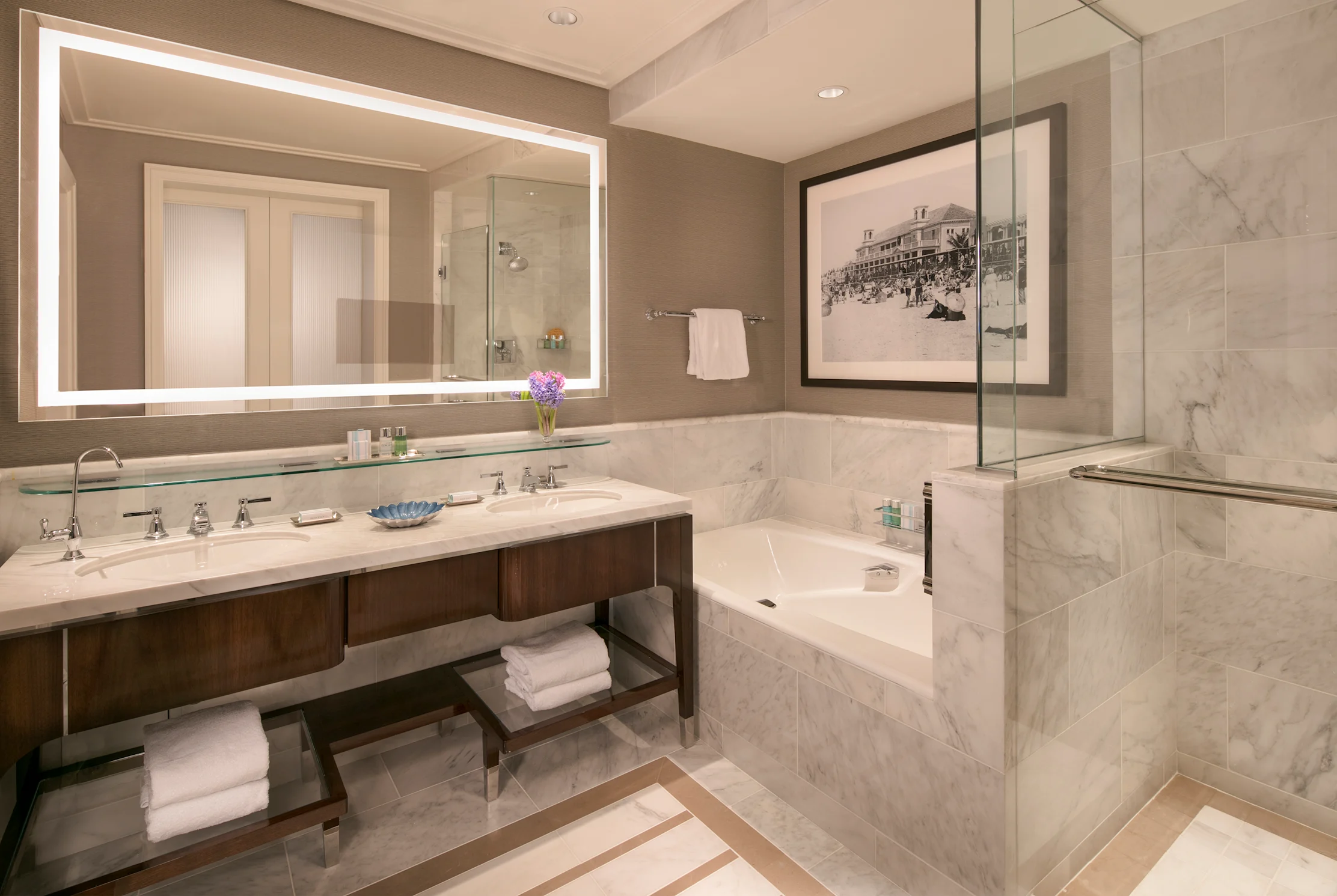 Luxury hotel bathroom with marble finishes, double vanity, backlit mirror, soaking tub, and glass shower.