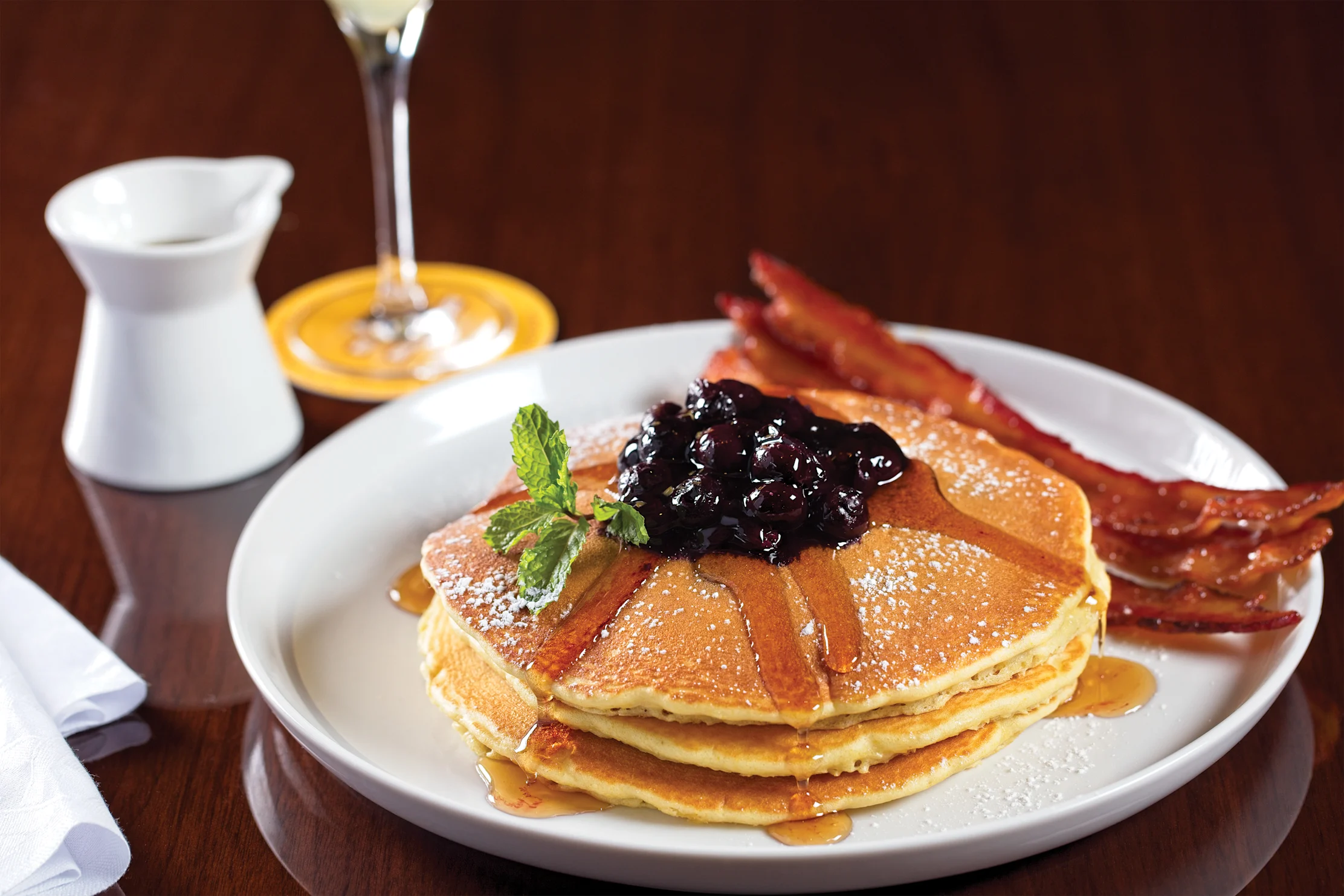 Stack of golden pancakes topped with blueberry compote, mint, and powdered sugar, served with bacon.