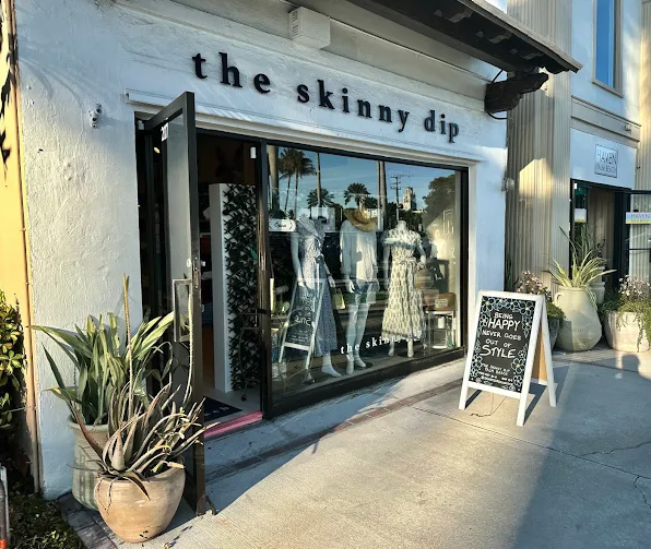 Storefront of "the skinny dip" boutique with white exterior, large display window showing mannequins, and plants flanking the entrance.