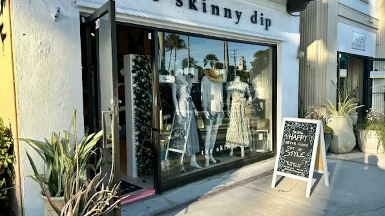 Storefront of "the skinny dip" boutique with white exterior, large display window showing mannequins, and plants flanking the entrance.