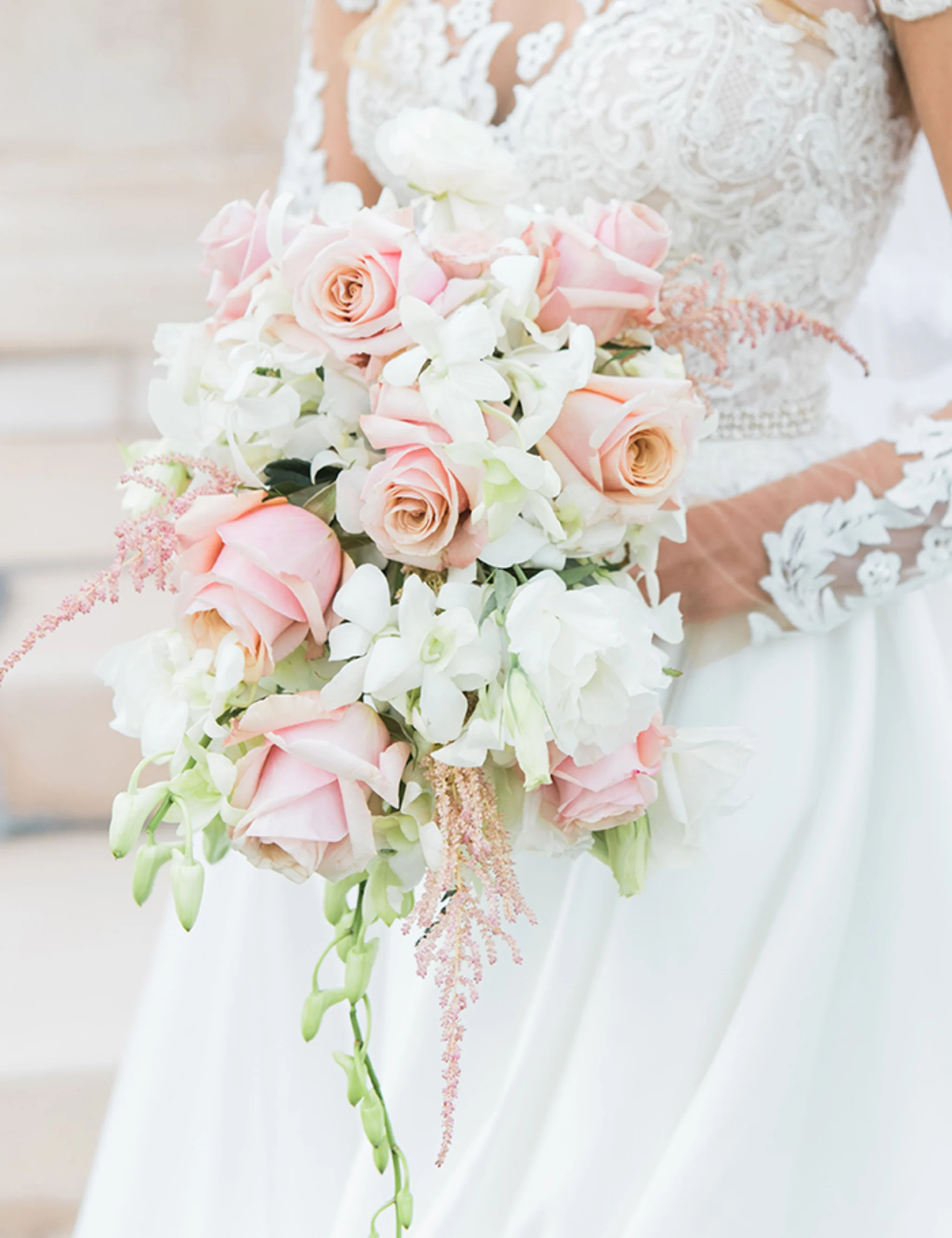Bride in white lace gown holding bouquet of pink roses and white flowers with trailing greenery