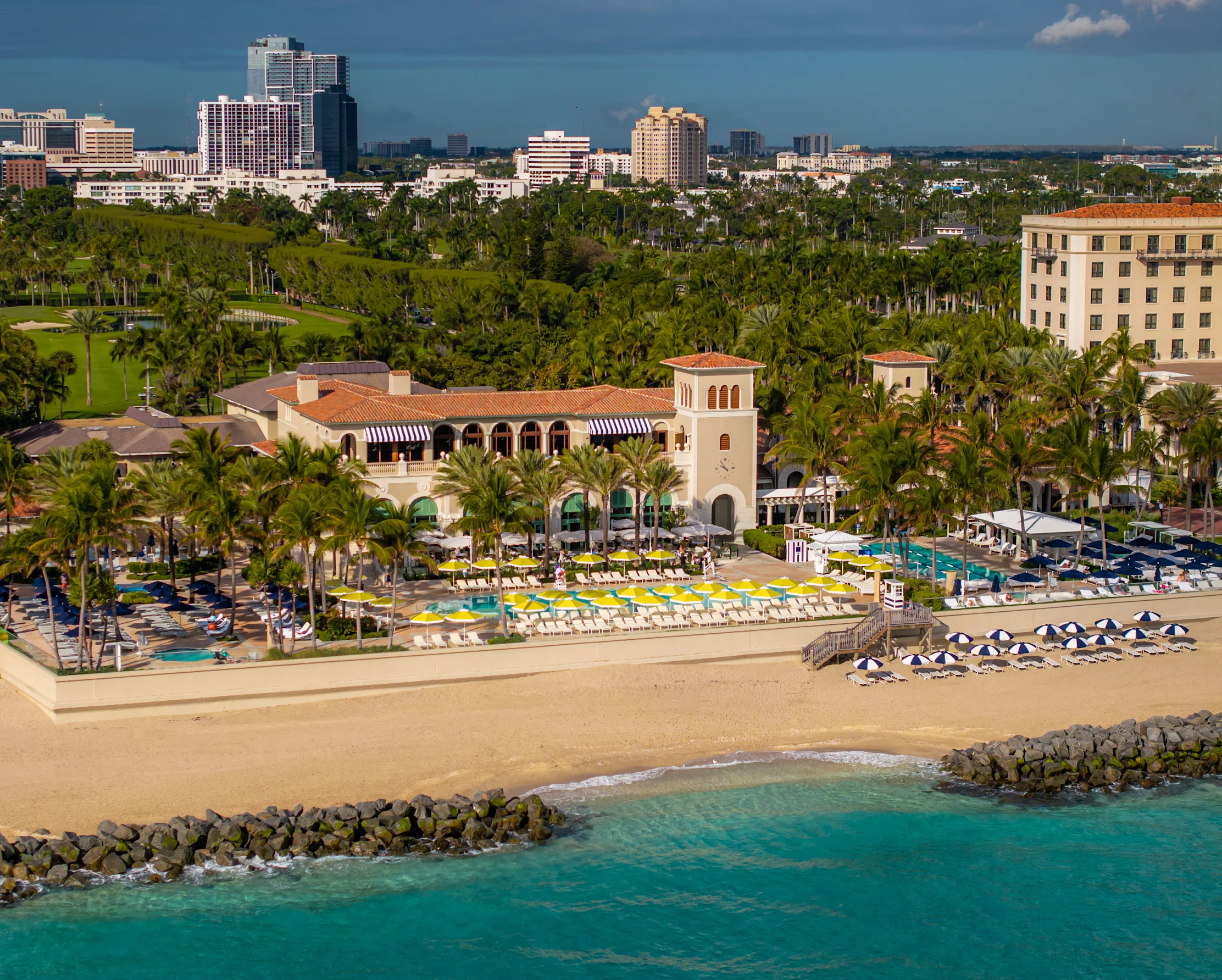Beachfront resort with Spanish-style architecture, yellow umbrellas, turquoise water, and city skyline.