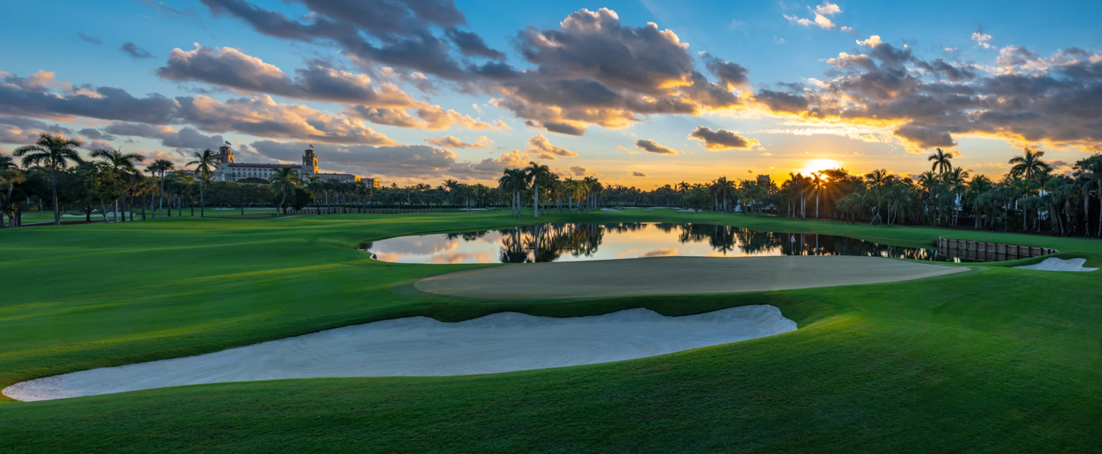 Golf course at sunset with palm trees, water hazard, sand bunker, and dramatic orange and blue sky.
