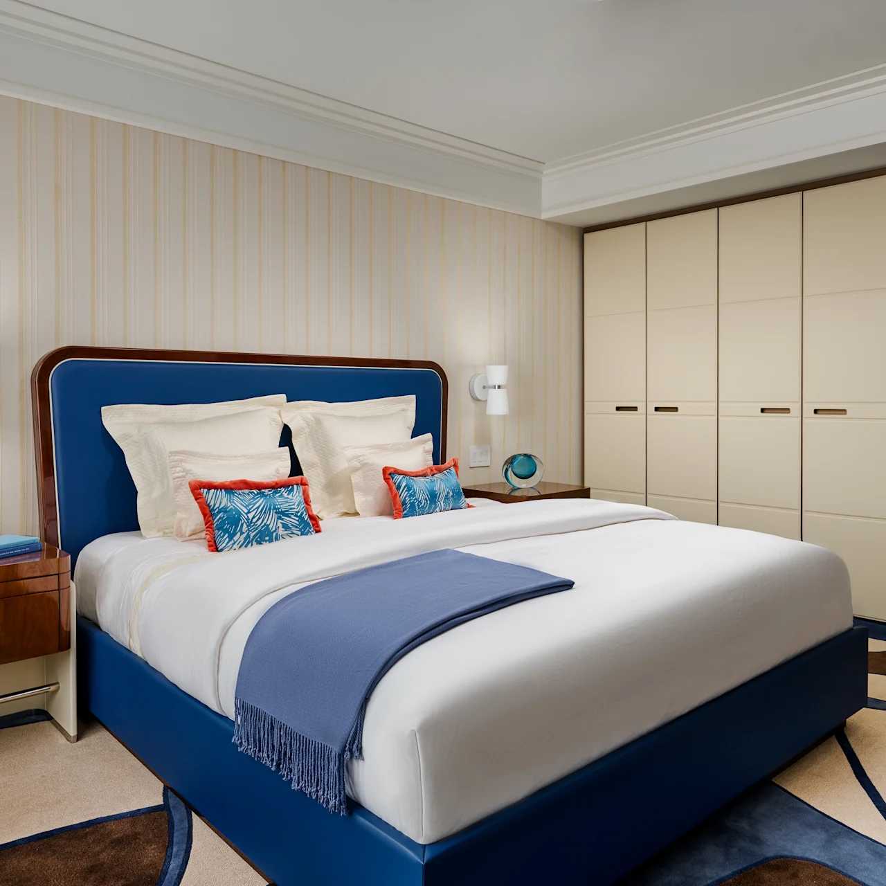 Modern bedroom with blue upholstered bed, white bedding, beige wardrobes, and striped wallpaper.