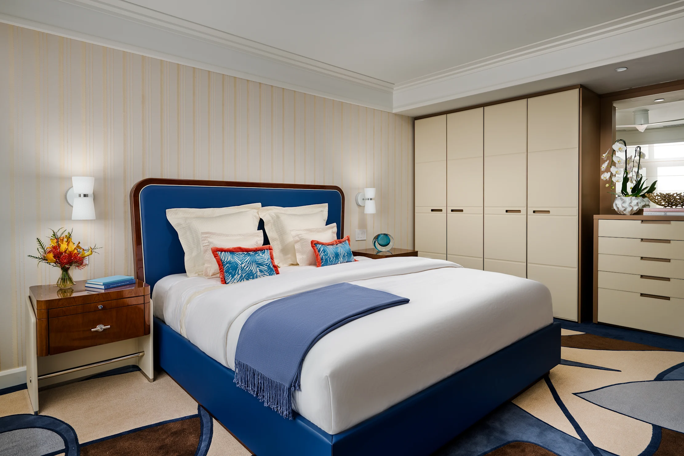Modern bedroom with blue upholstered bed, white bedding, beige wardrobes, and striped wallpaper.